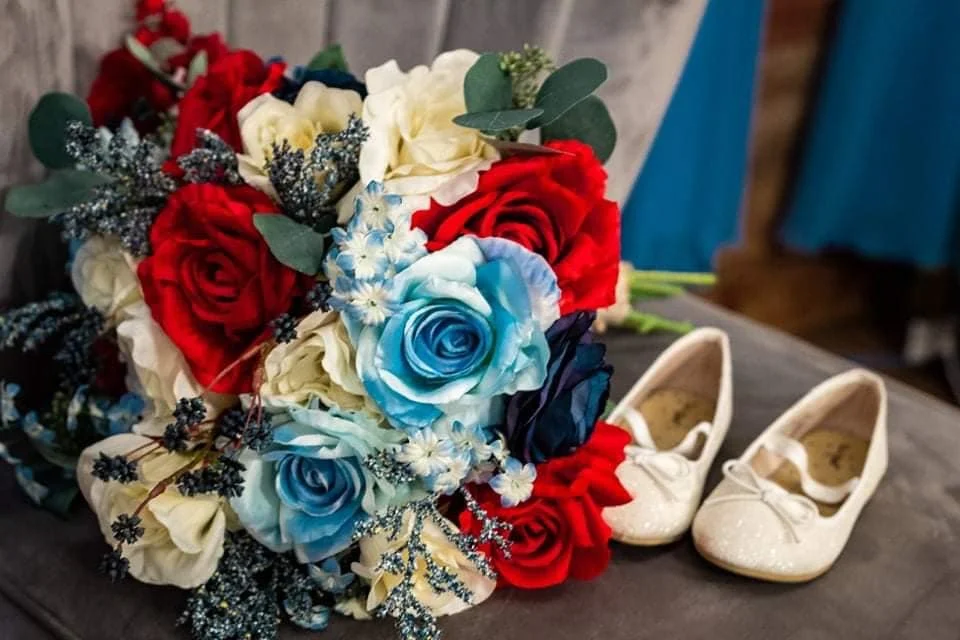 A bouquet of artificial flowers including red, white, and blue roses, with greenery, placed on a dark surface. Next to the bouquet are a pair of white children's shoes with lace details and bows.