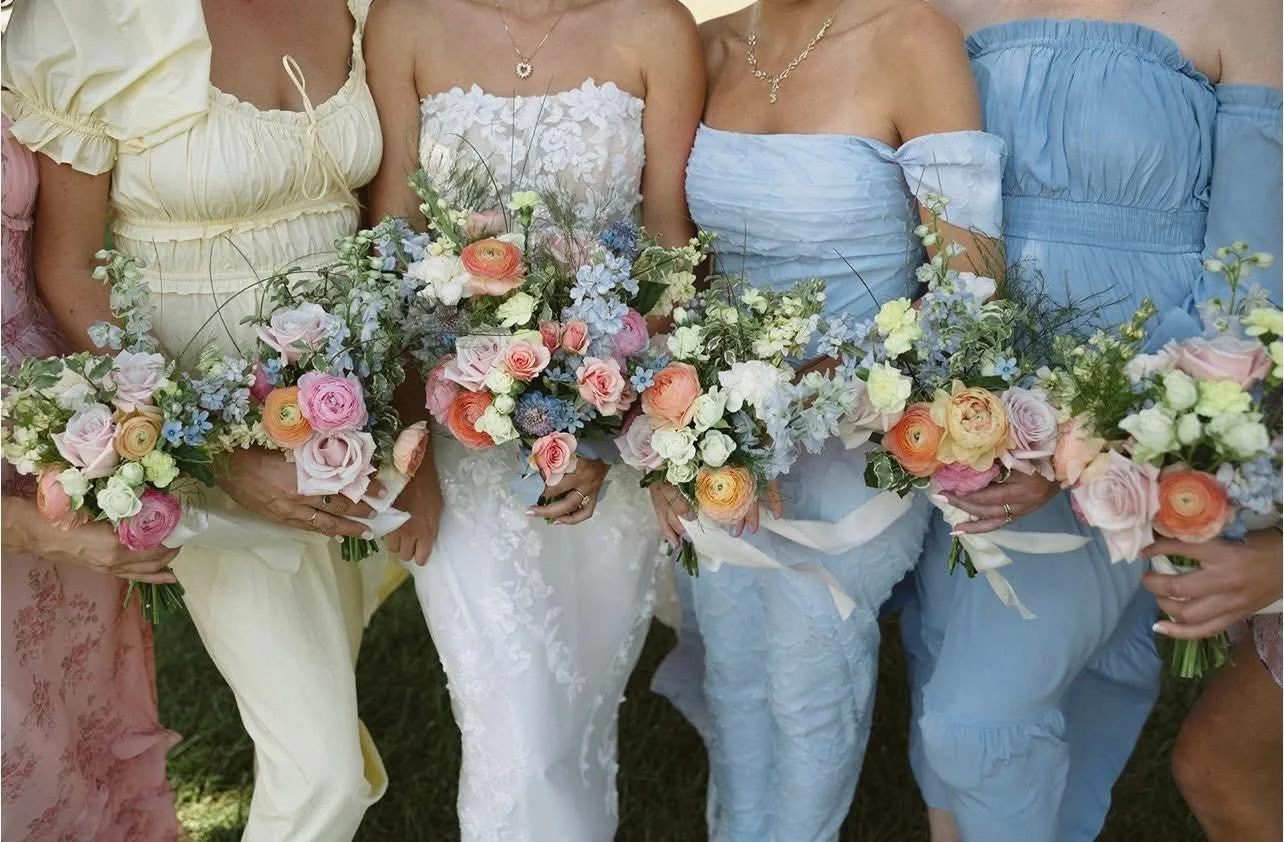 Group of women holding colorful flower bouquets, wearing pastel dresses, possibly at a wedding or celebration.