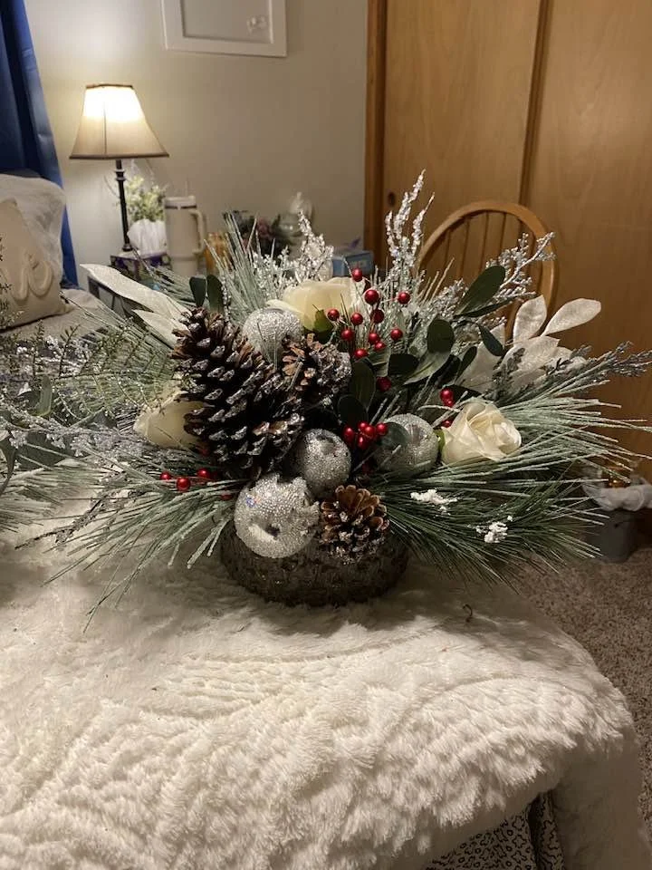 A festive Christmas floral arrangement with pine cones, silver ornaments, white roses, and red berries in a dark vessel, placed on a cream-colored fluffy tablecloth in a cozy room.