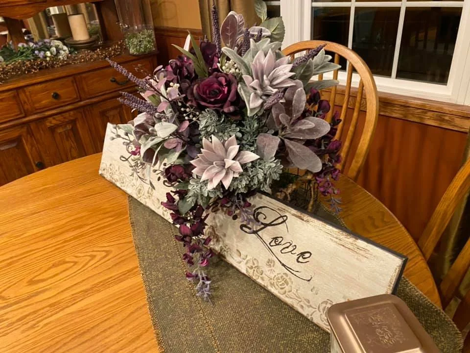 Decorative floral arrangement in a white wooden box with the word 'Love' written on it, sitting on a wooden dining table.