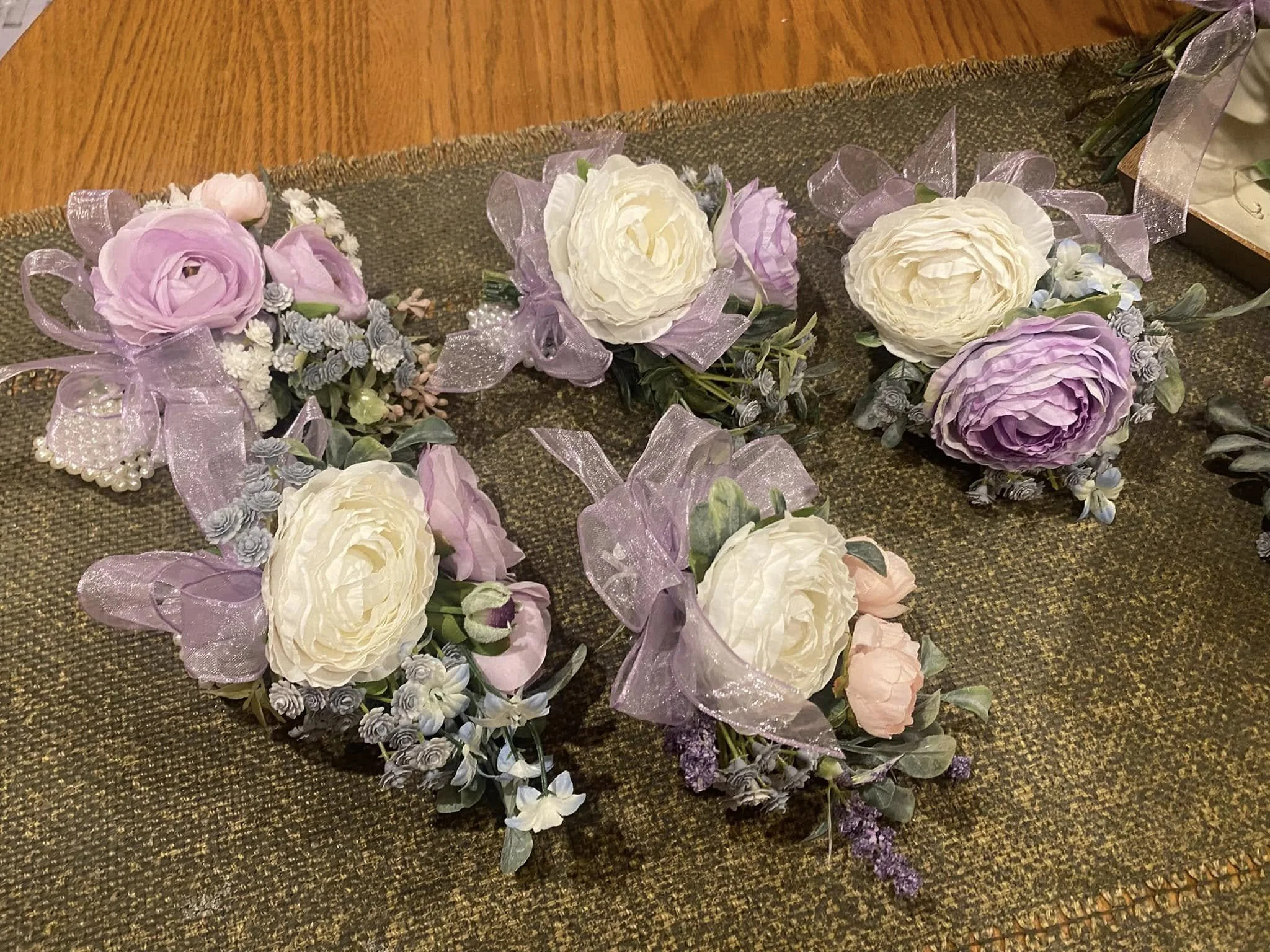 Four floral corsages with white and purple flowers, lavender ribbons, and pearl accents on a textured gold and brown surface.