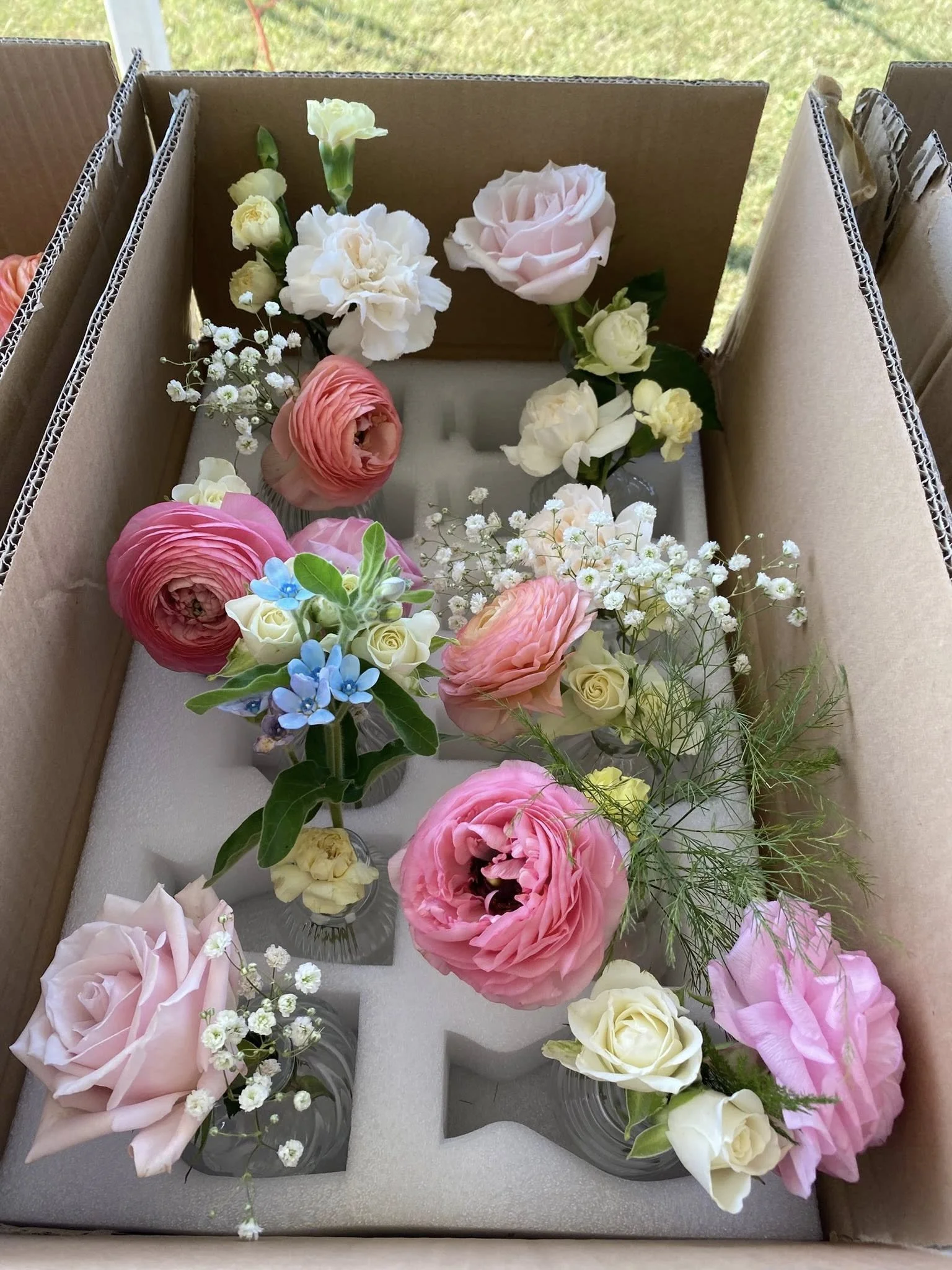 Assorted fresh flowers in small glass vases, including roses, ranunculus, and baby's breath, arranged in a cardboard box with foam padding.