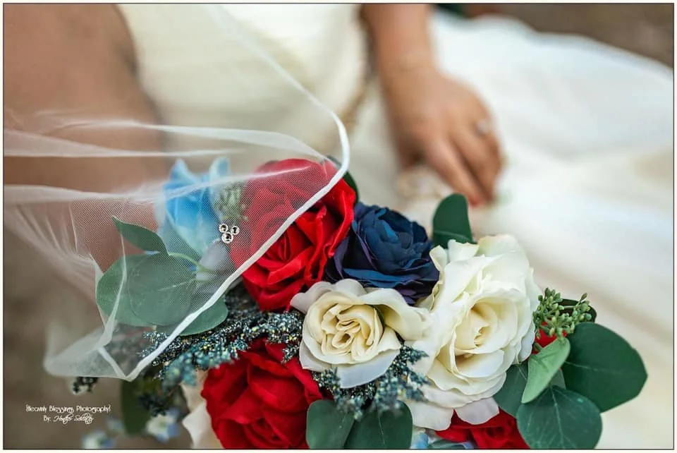 A wedding bouquet with red, white, and blue roses, green leaves, and a decorative veil.