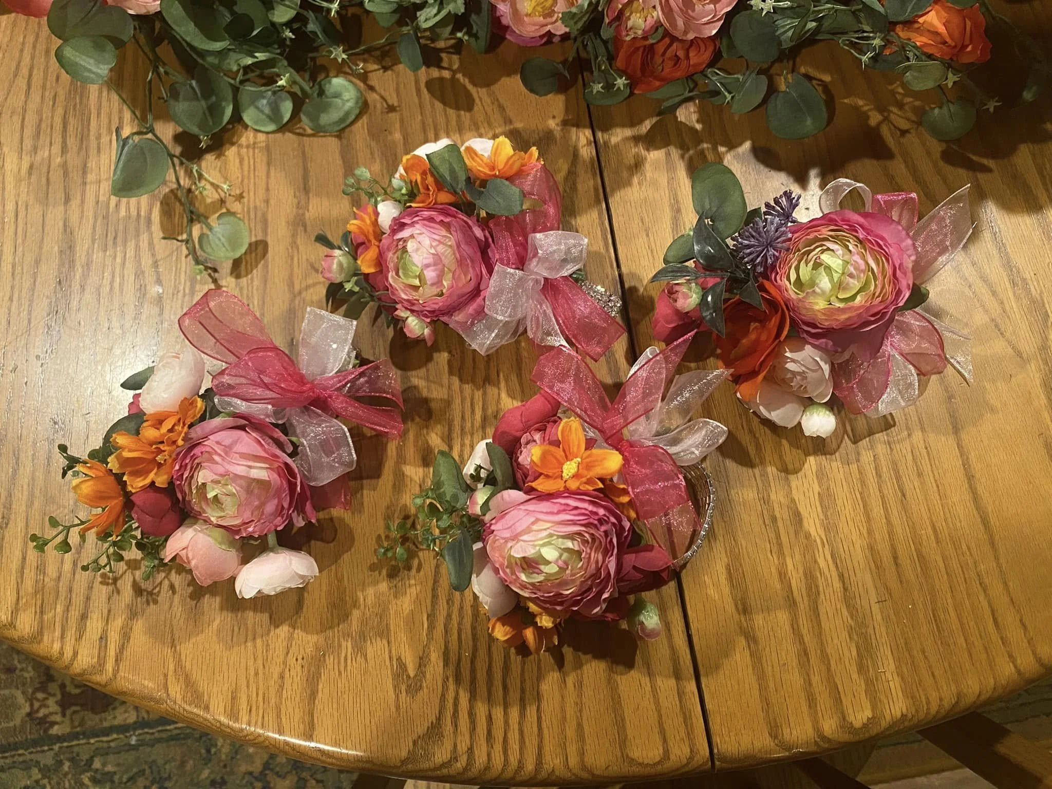 Corsages with pink peonies, orange daisies, purple thistles, green leaves, and pink and red sheer ribbons.
