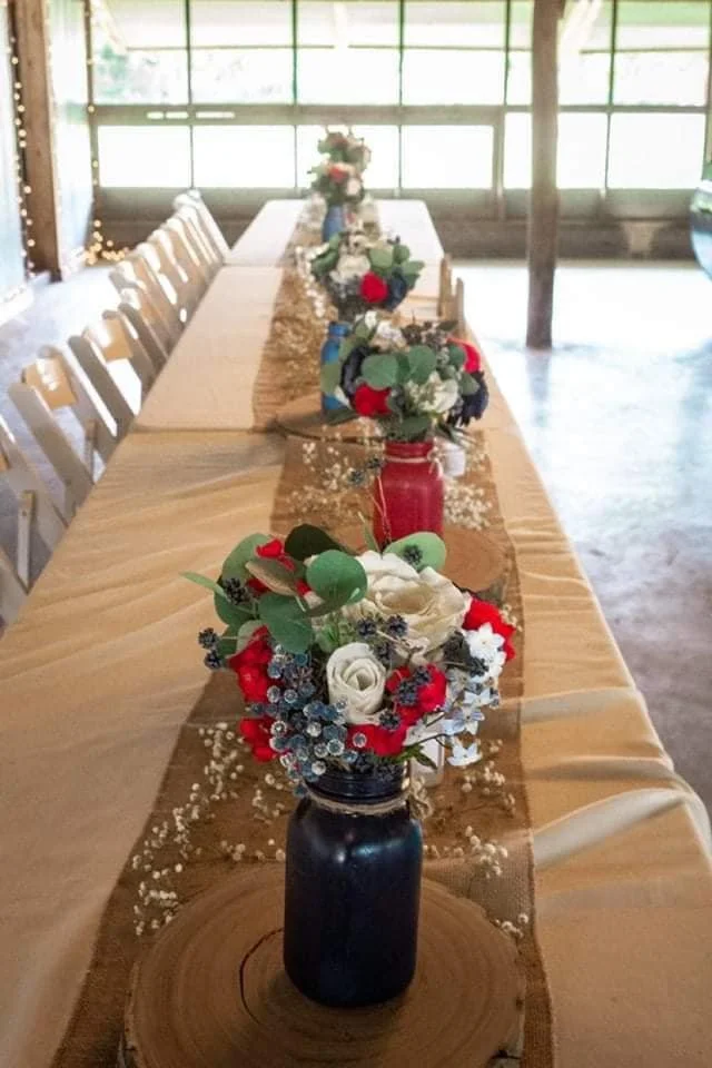 Long wooden table decorated with multiple floral arrangements in black, red, green, and white vases, with a beige table runner and scattered baby's breath flowers, set in a spacious, well-lit room with large windows.