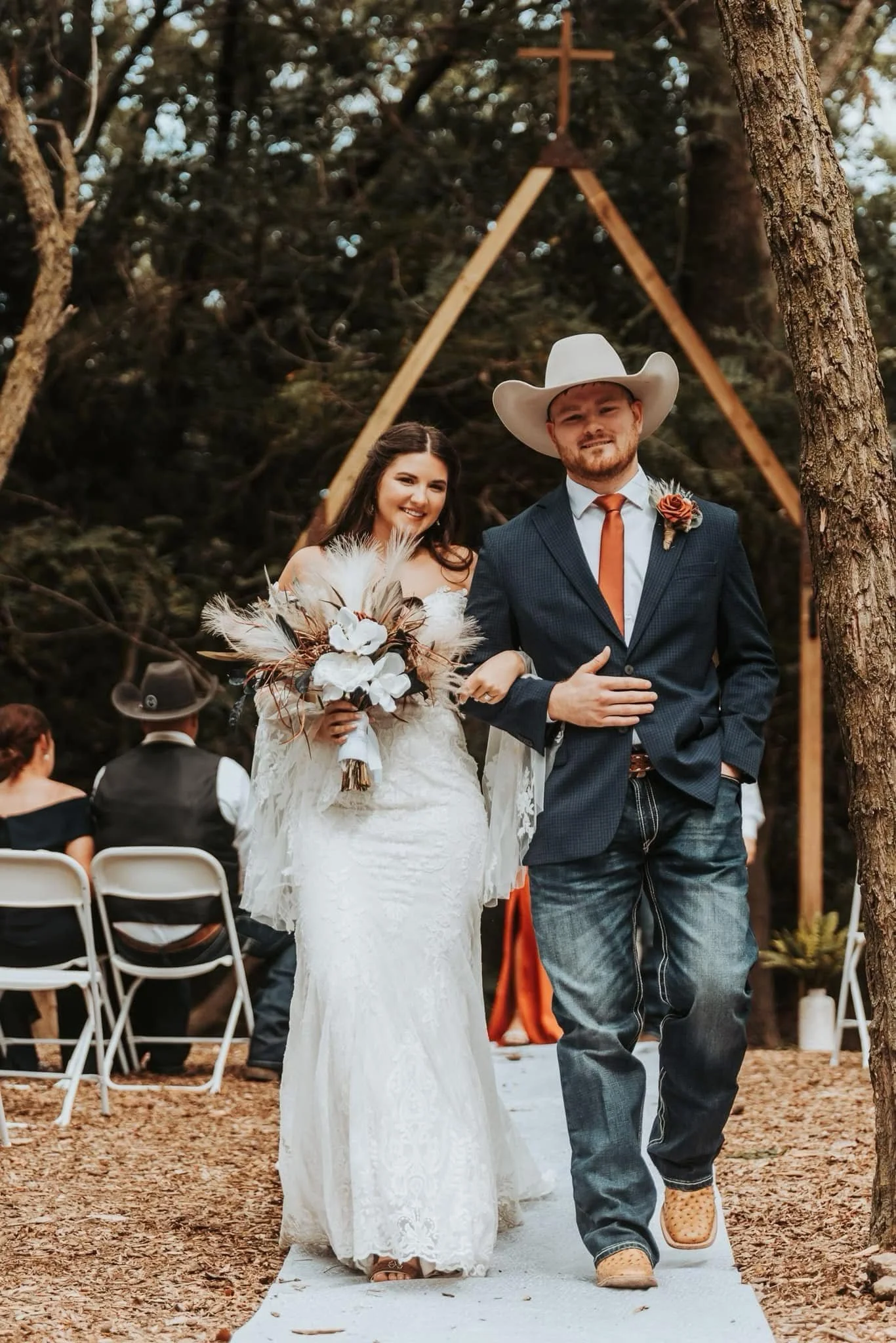 A bride and groom walking down the aisle during an outdoor wedding ceremony. The bride holds a bouquet, wearing a white lace dress. The groom wears a dark suit with a white cowboy hat and a orange tie.