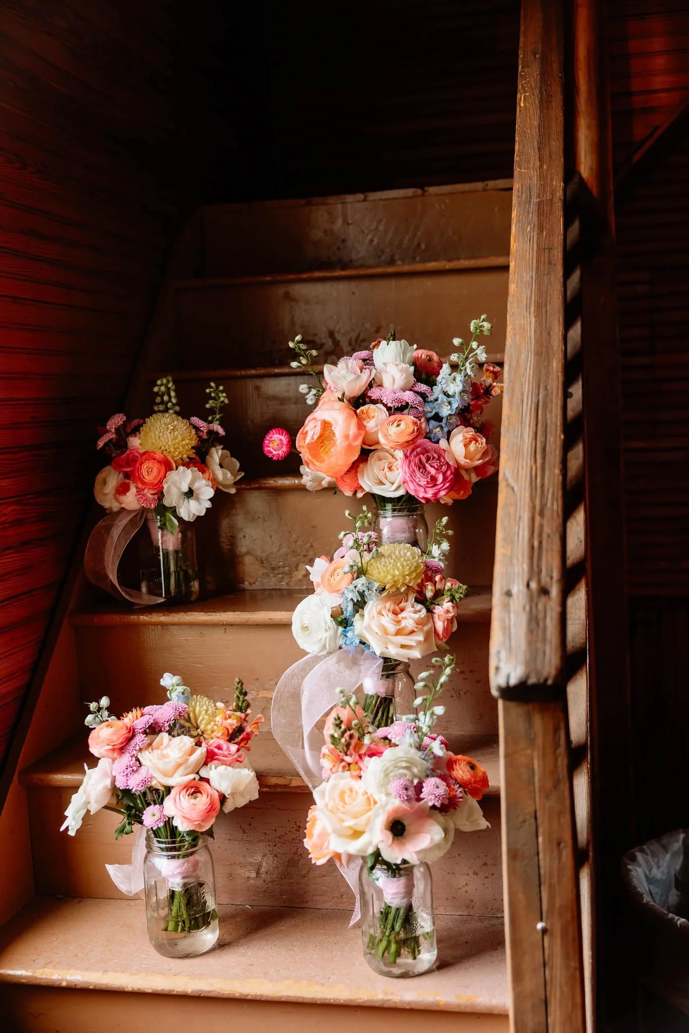 Multiple bouquets of pink, peach, white, and purple flowers in glass jars positioned on a worn wooden staircase.