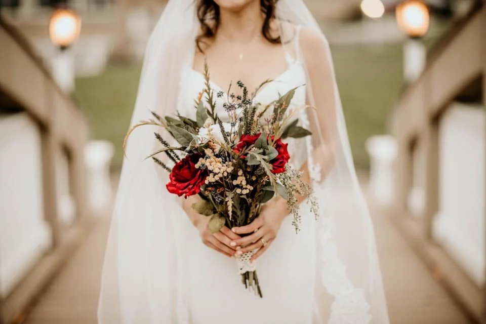 Bride holding a bouquet of red roses and assorted flowers on a wooden walkway.