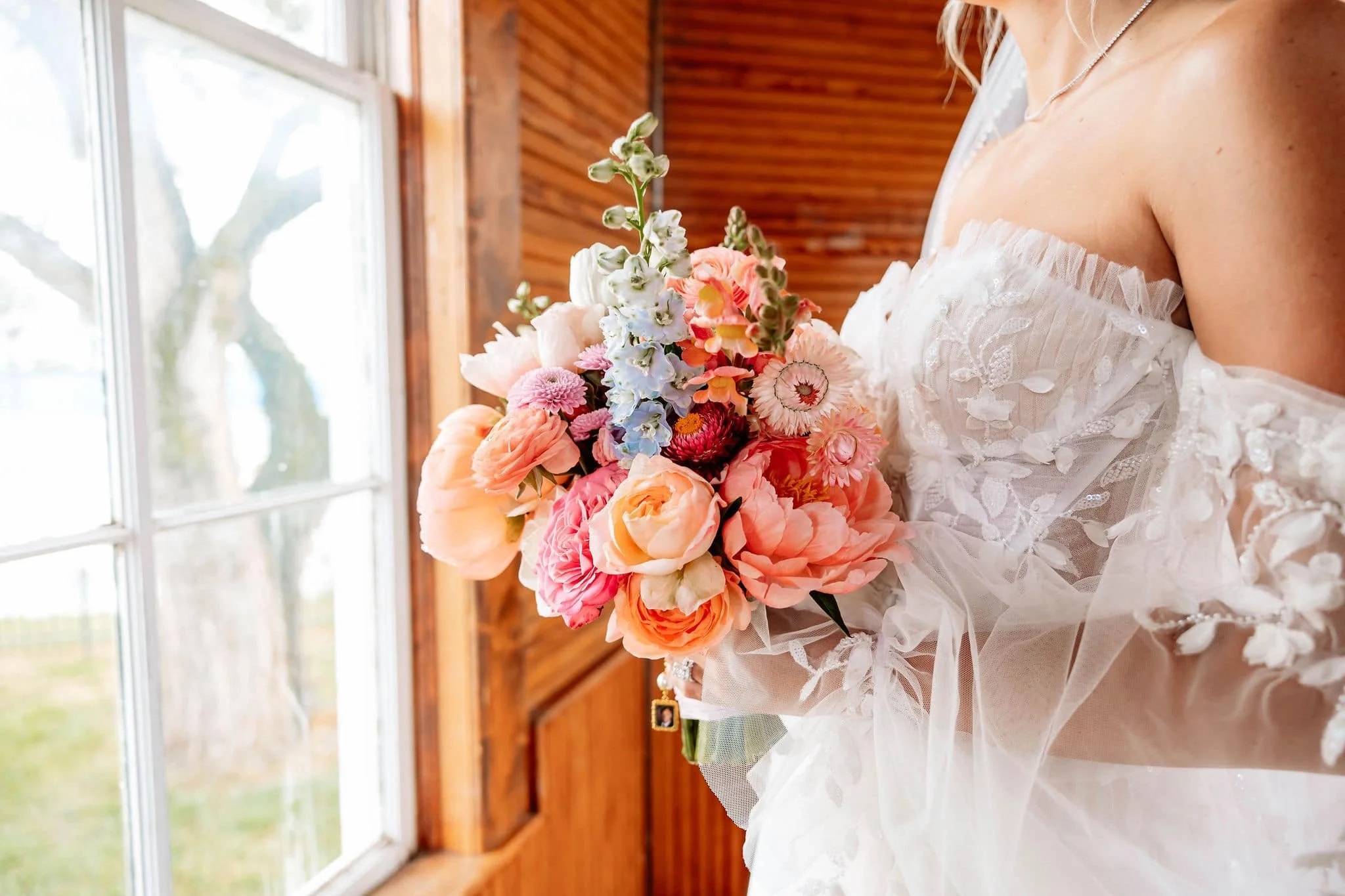 A bride holding a bouquet of pink, white, and light blue flowers inside a wooden room near a window.