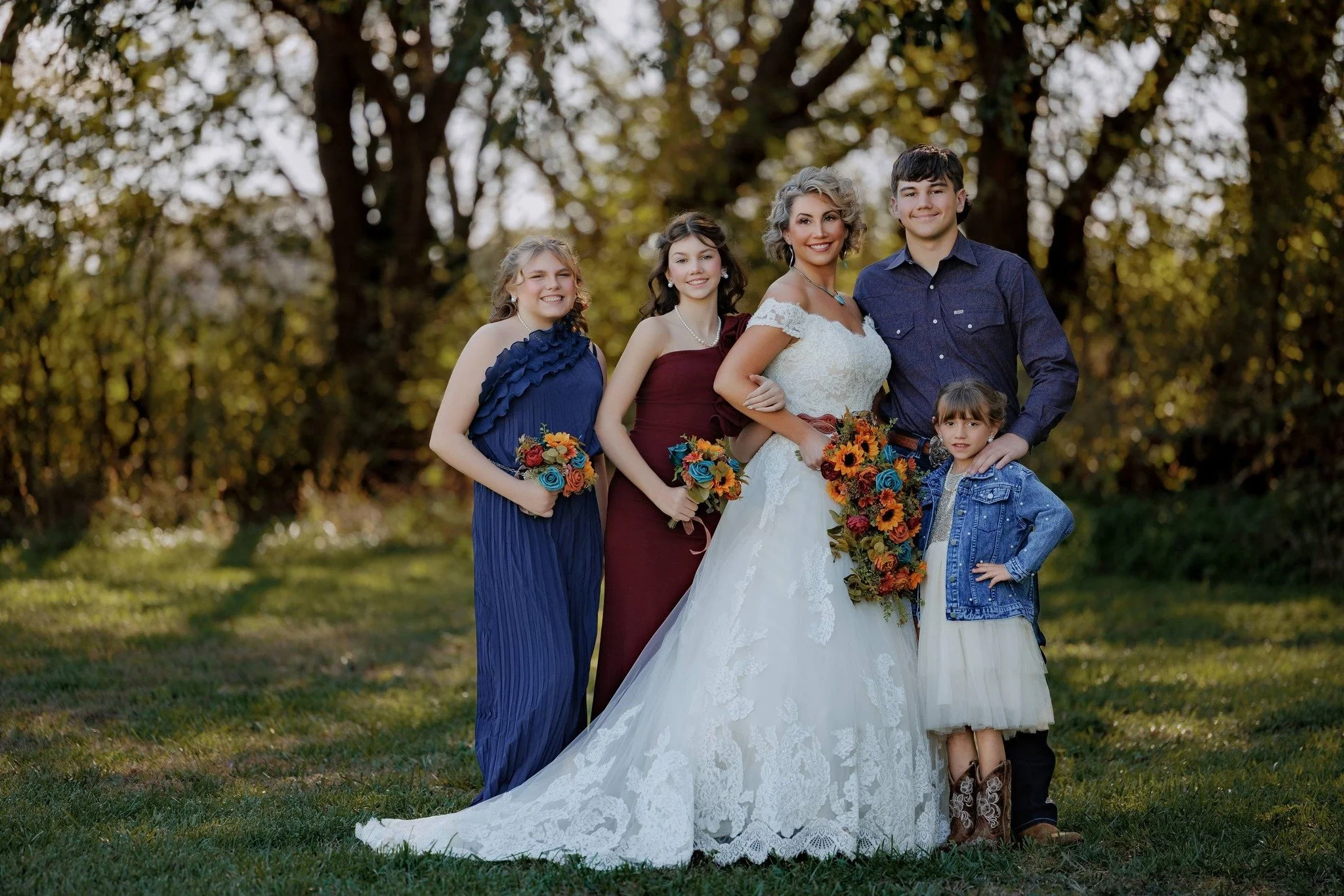 A group of five people, including a bride in a white wedding gown holding a bouquet, posing outdoors on a grassy area with trees in the background during daytime.