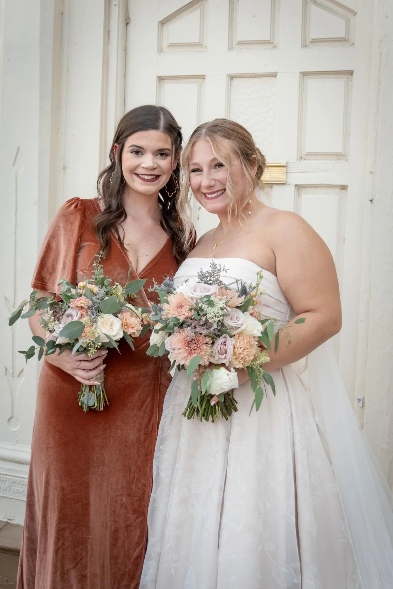 Two women smiling and holding bouquets of flowers, standing in front of a white door.