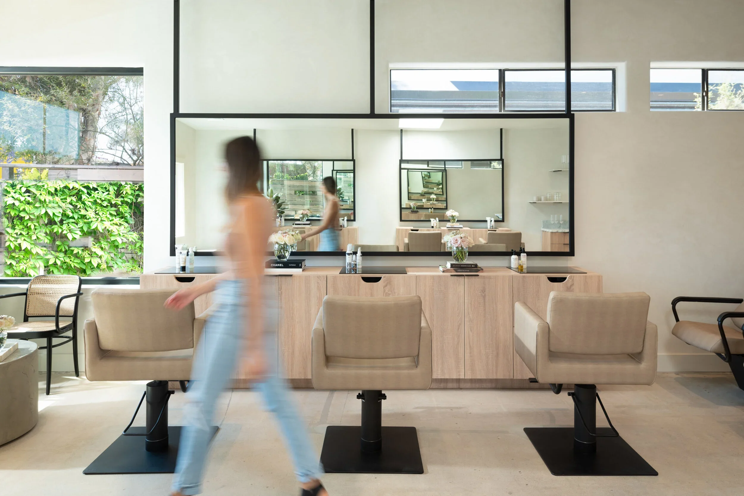 Modern hair salon with beige chairs and large mirrors, a woman walking past, and natural light coming through windows.