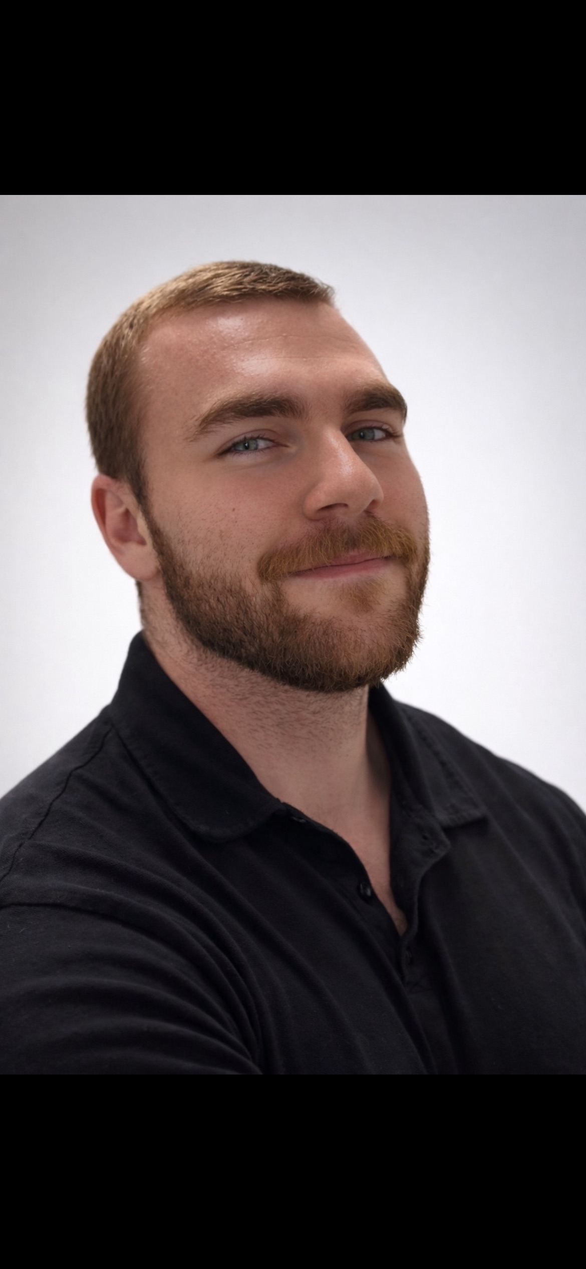 Portrait of a bearded man with short hair, blue eyes, and a black collared shirt, smiling slightly against a plain light background.