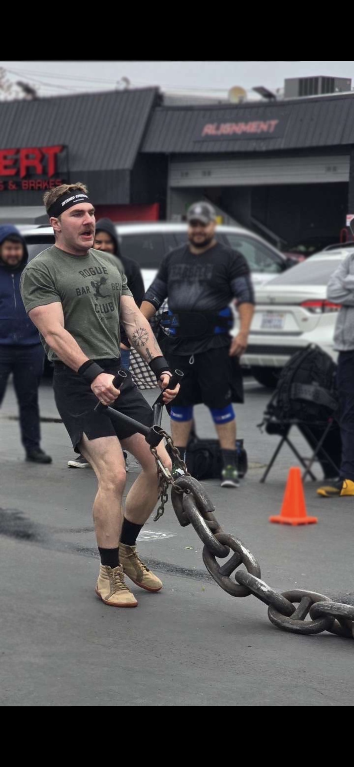 Man in workout clothes pulling a large heavy chain during a strongman competition, with others and cars in the background.
