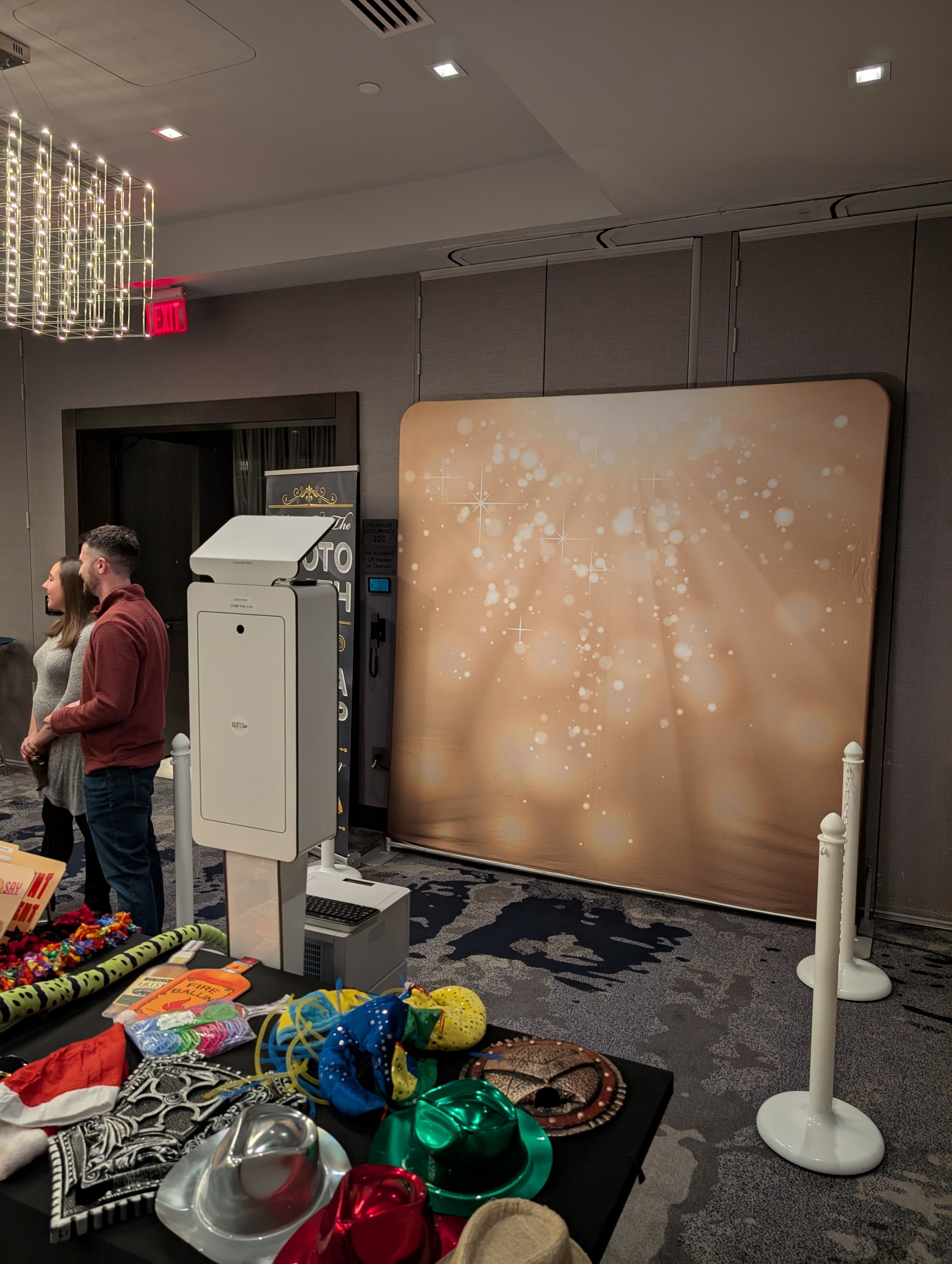 Indoor event space with a photo booth backdrop featuring bokeh lights, people standing near a table with colorful accessories, and various items on display.