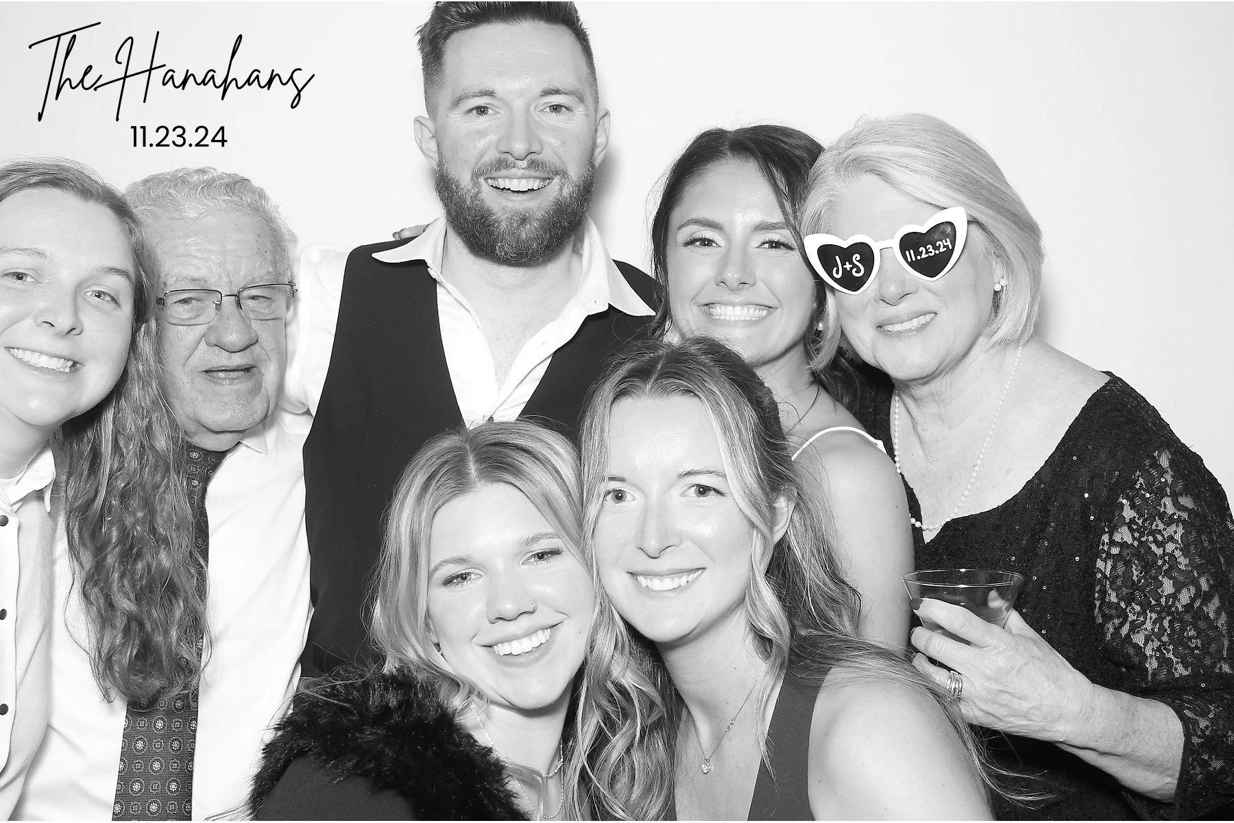 Group of seven people taking a black and white photo at a celebration event, with a woman wearing novelty sunglasses that say 'J + S' and a date, holding a drink, and the event name 'The Hanahans' along with the date 11.23.24 written in the top left corner.