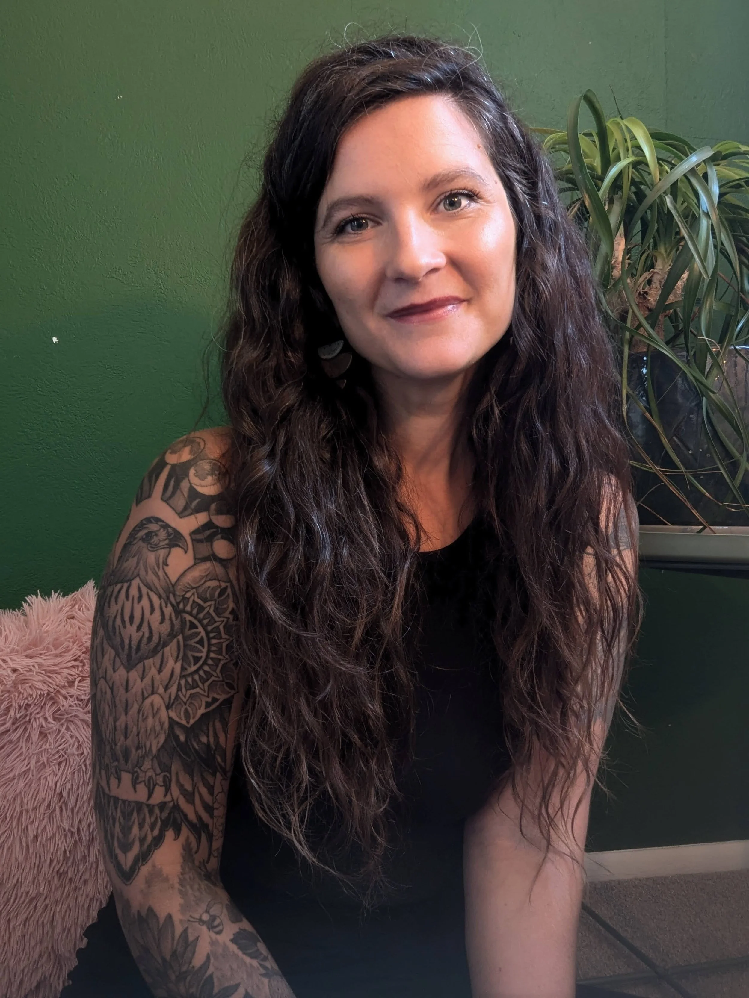 A woman with long, curly dark hair sitting on a pink furry chair in front of a green wall, with a large potted plant behind her, wearing a sleeveless black top and earrings, smiling at the camera.