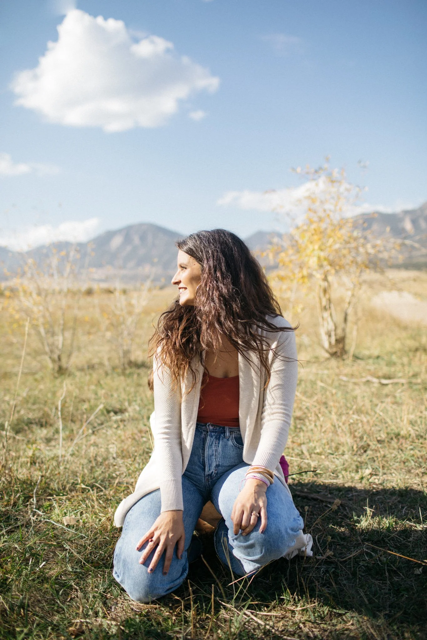 A woman with long, curly hair smiling and sitting on the grass in a field during daytime with mountains in the background under a partly cloudy sky.