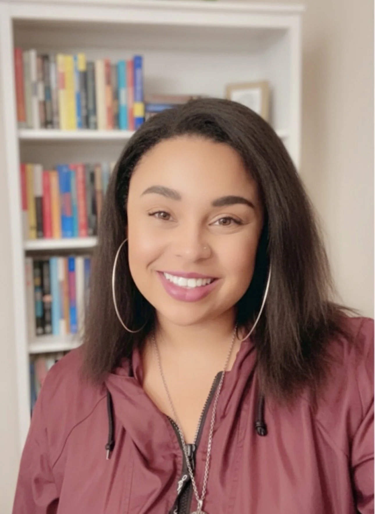 A smiling woman with long dark hair, hoop earrings, wearing a maroon jacket, standing in front of a bookshelf filled with various books.