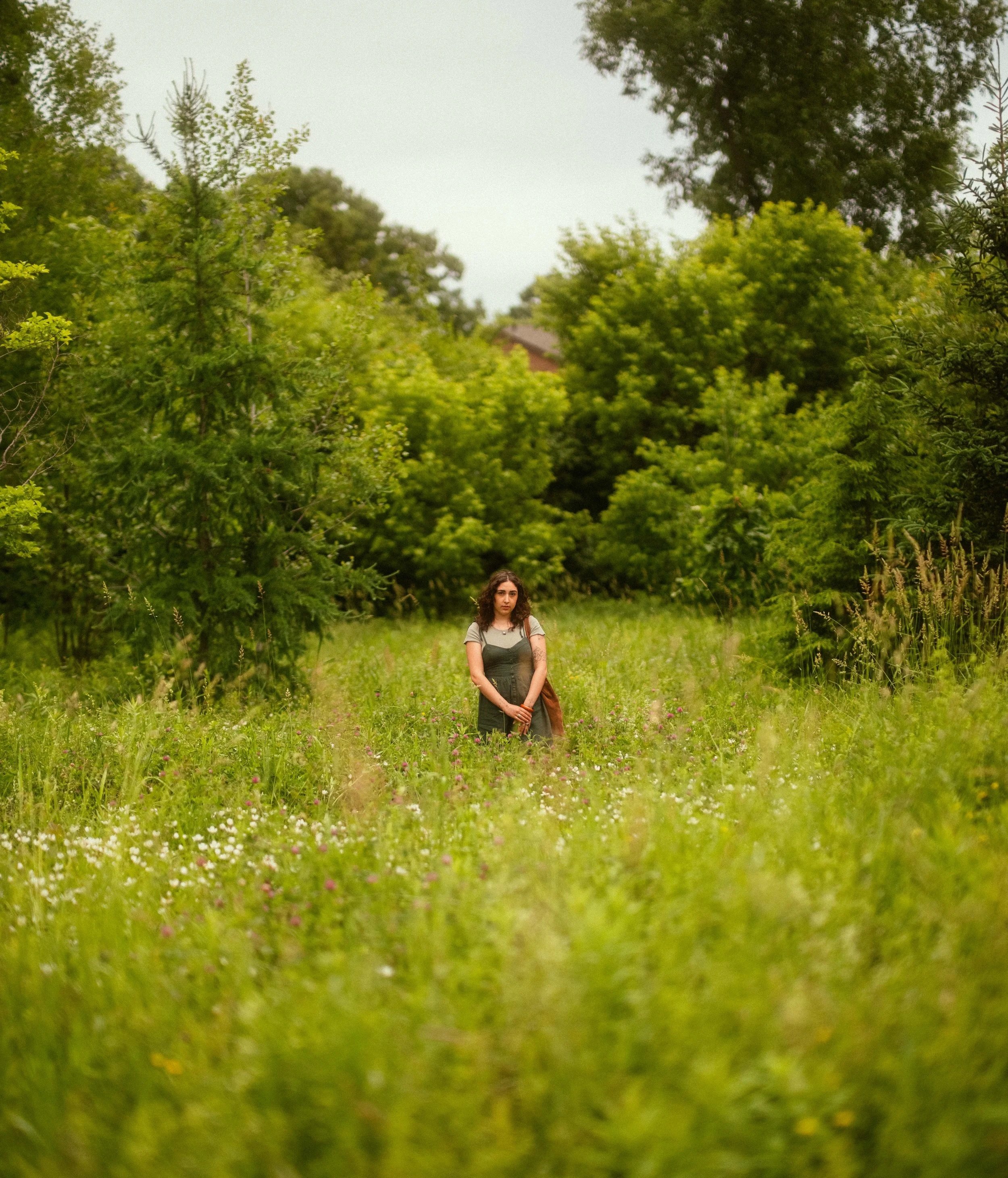 A woman standing in a lush green meadow with tall grass and wildflowers, surrounded by trees and foliage on a cloudy day.