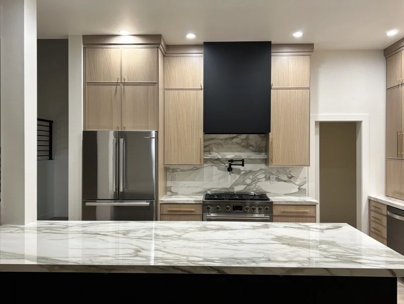 Modern kitchen with light wood cabinets, stainless steel refrigerator, marble backsplash and countertops, black range hood, and stainless stove, with a large marble island in the foreground.