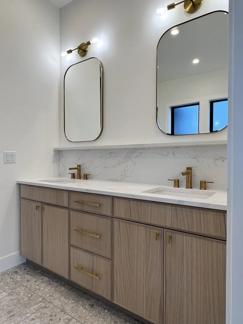 Modern bathroom vanity with a dual sink, gold fixtures, light wood cabinetry, marble countertop, and two large mirrors above. There are two wall-mounted light fixtures above each mirror and a window reflected in one mirror.