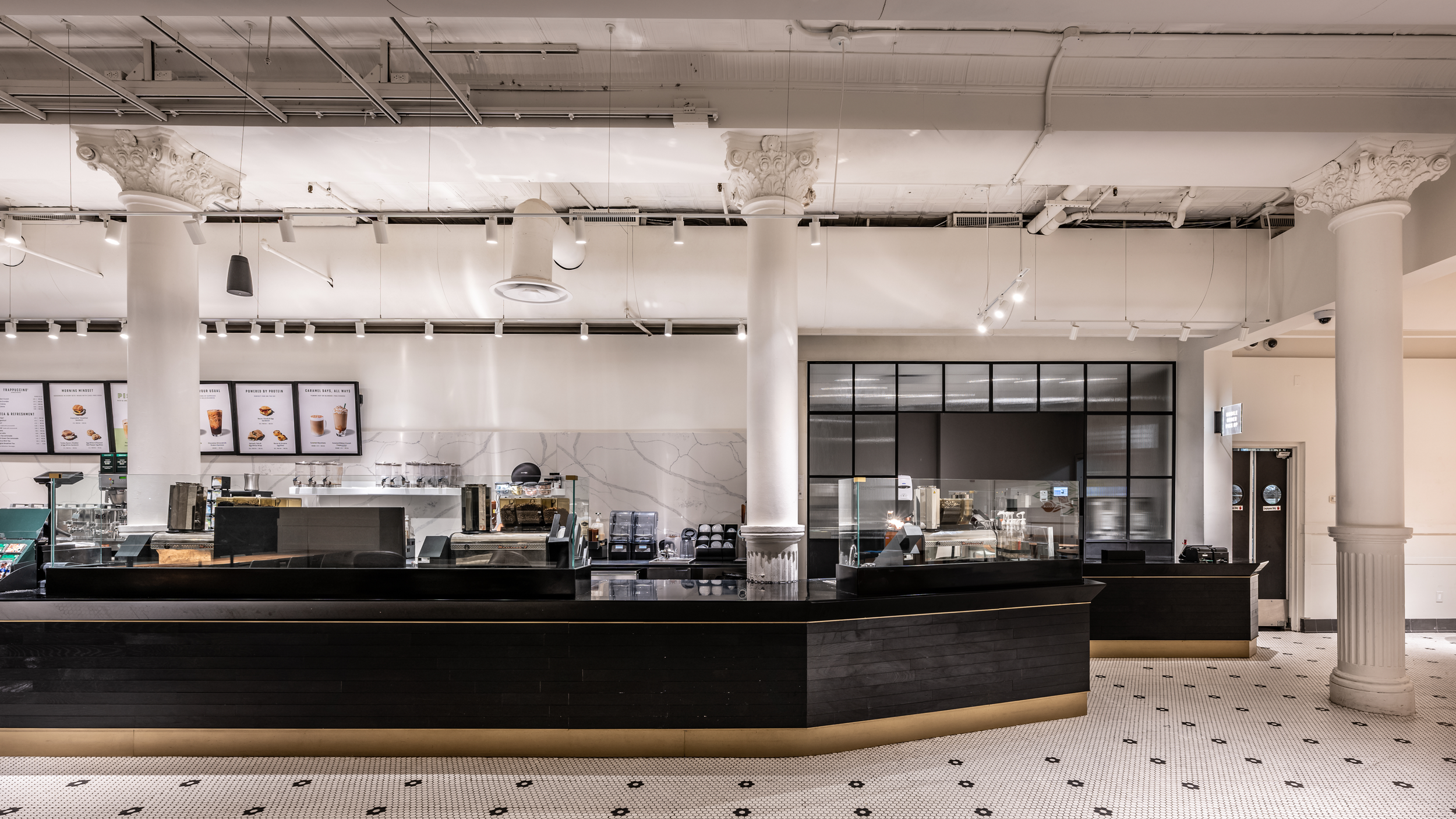 Interior of a modern coffee shop with a black counter, white marble walls, and high ceilings with decorative columns and hanging lights.