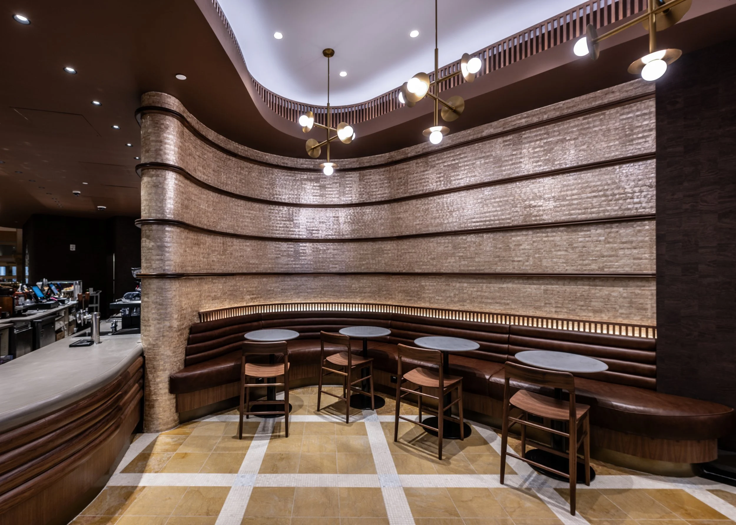 Interior of a modern cafe with a curved brown leather seating area, small round tables, and wooden chairs. The wall behind features horizontal textured light-colored tiles with dark accent lines. The ceiling has contemporary light fixtures with round