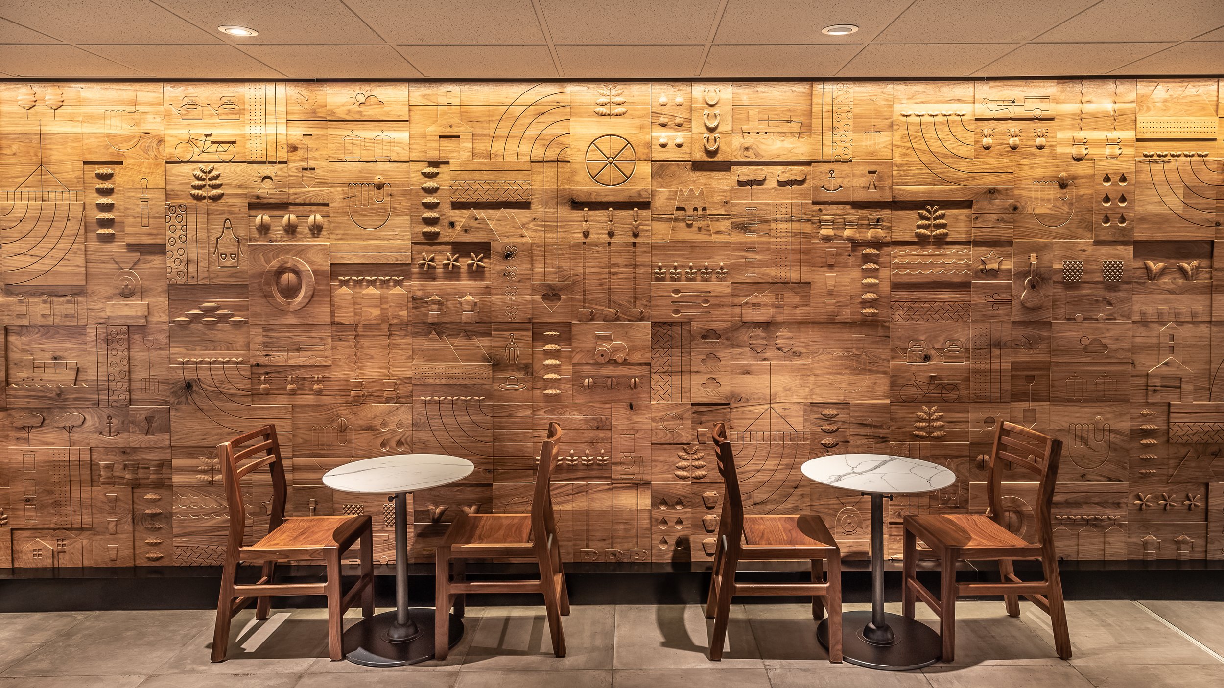 A wooden wall with intricate carved patterns and symbols in a modern cafe, with two round tables and four wooden chairs in front.