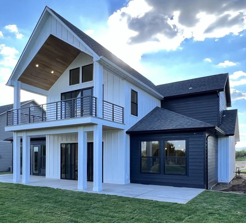 Modern two-story house with white and dark blue exterior siding, large windows, a small balcony, and a well-maintained lawn under a partly cloudy sky.