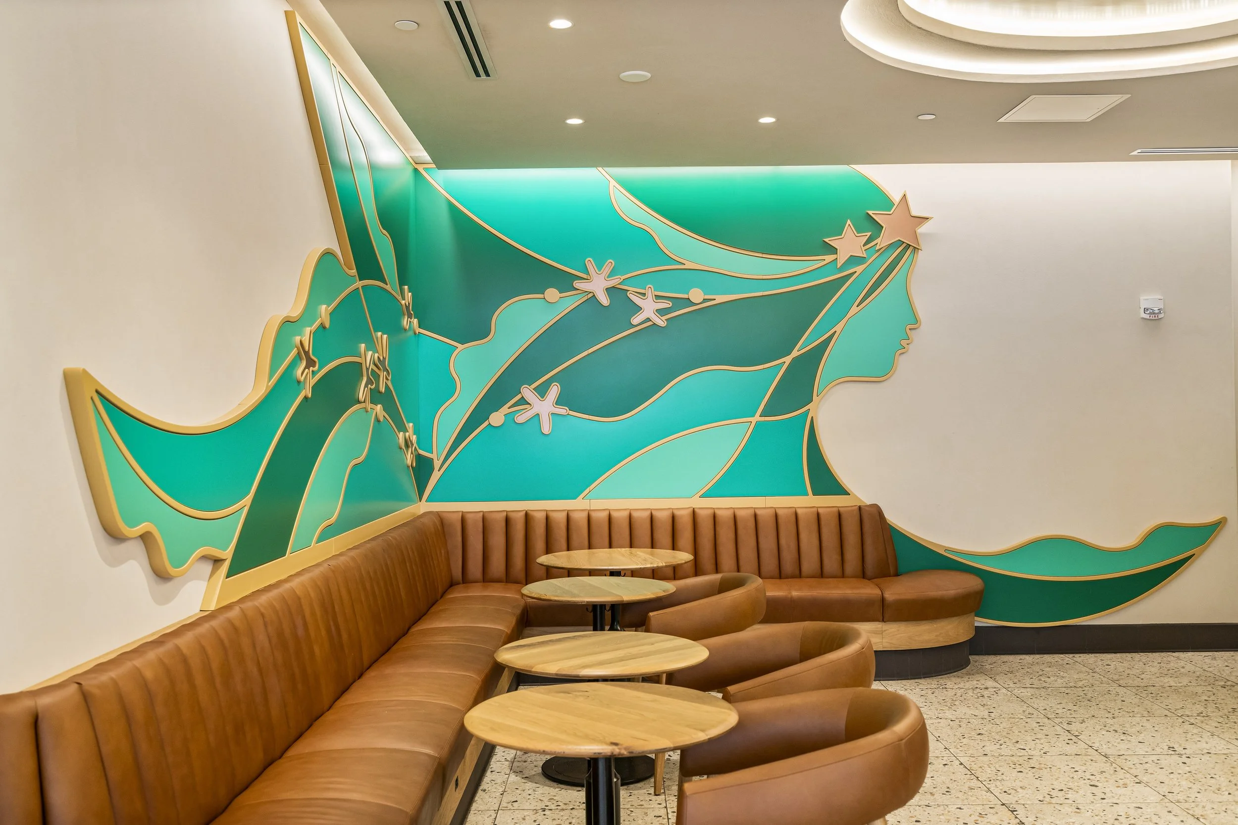 Interior of a modern cafe with a decorative wall mural of a stylized woman's profile with flowing hair, stars, and abstract lines in teal and gold. Brown leather seating and wooden tables arranged in front of the mural.