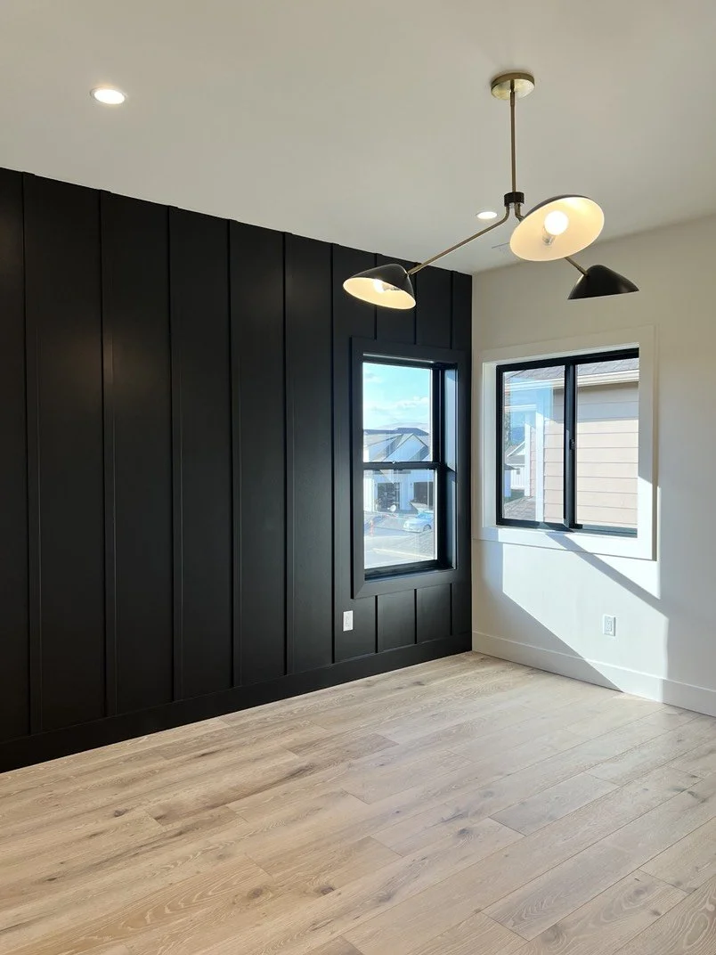 Empty room with black paneled accent wall, two black-framed windows, light wood flooring, and modern ceiling light fixture with rotating arms and multiple black and white lamps