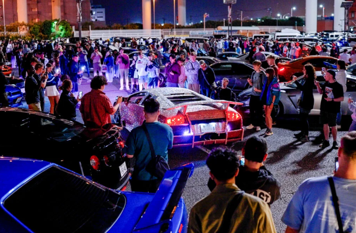 Crowd of car enthusiasts gathered around a modified sports car at night, at a car meet or event.