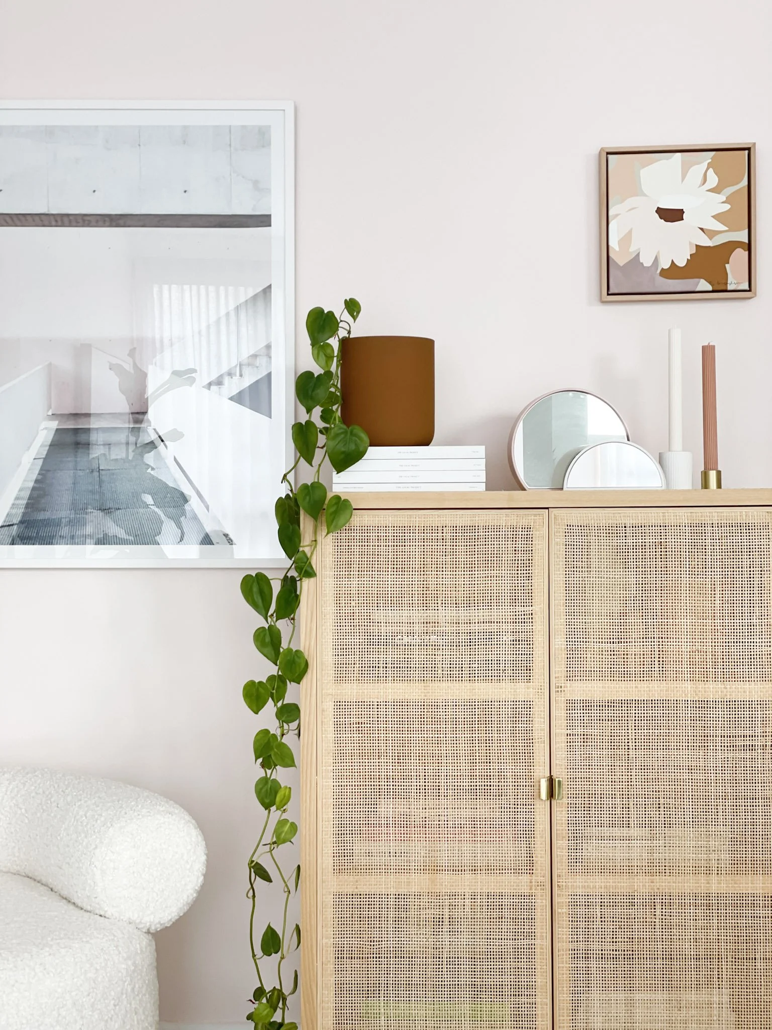 A cozy corner of a living room with a cream armchair, a wooden cabinet with a rattan front, a potted heart-leaf philodendron plant, and decorative items including round mirrors, a brown lamp, and candles on top of the cabinet. There is a framed art p