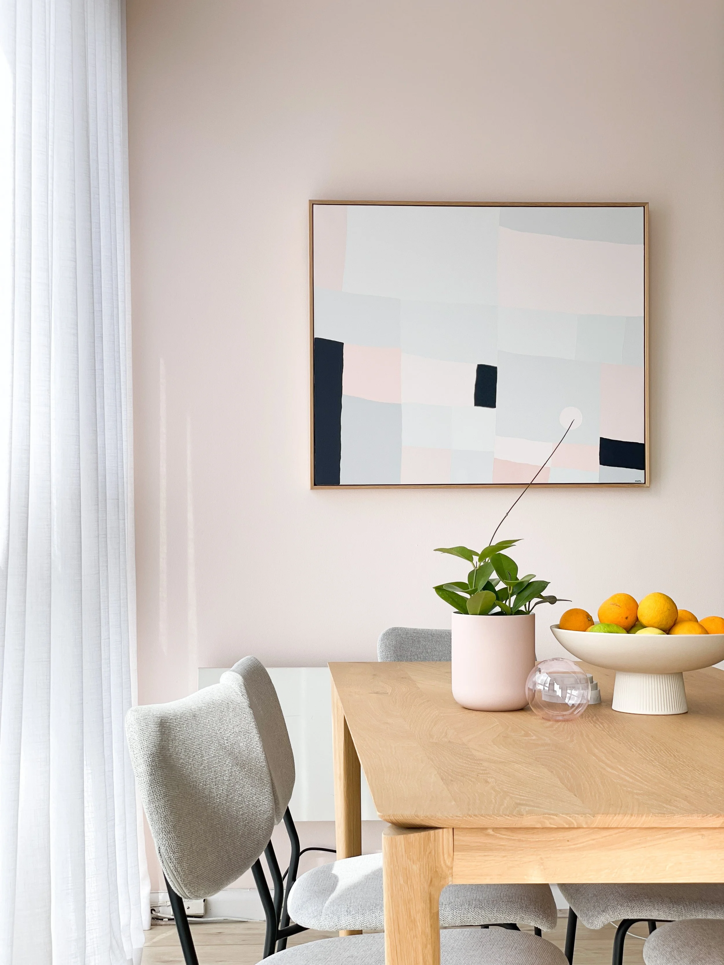 Minimalist dining room with a wooden table, gray upholstered chairs, a potted plant, a bowl of citrus fruits, a glass decorative object, and abstract artwork on a beige wall.