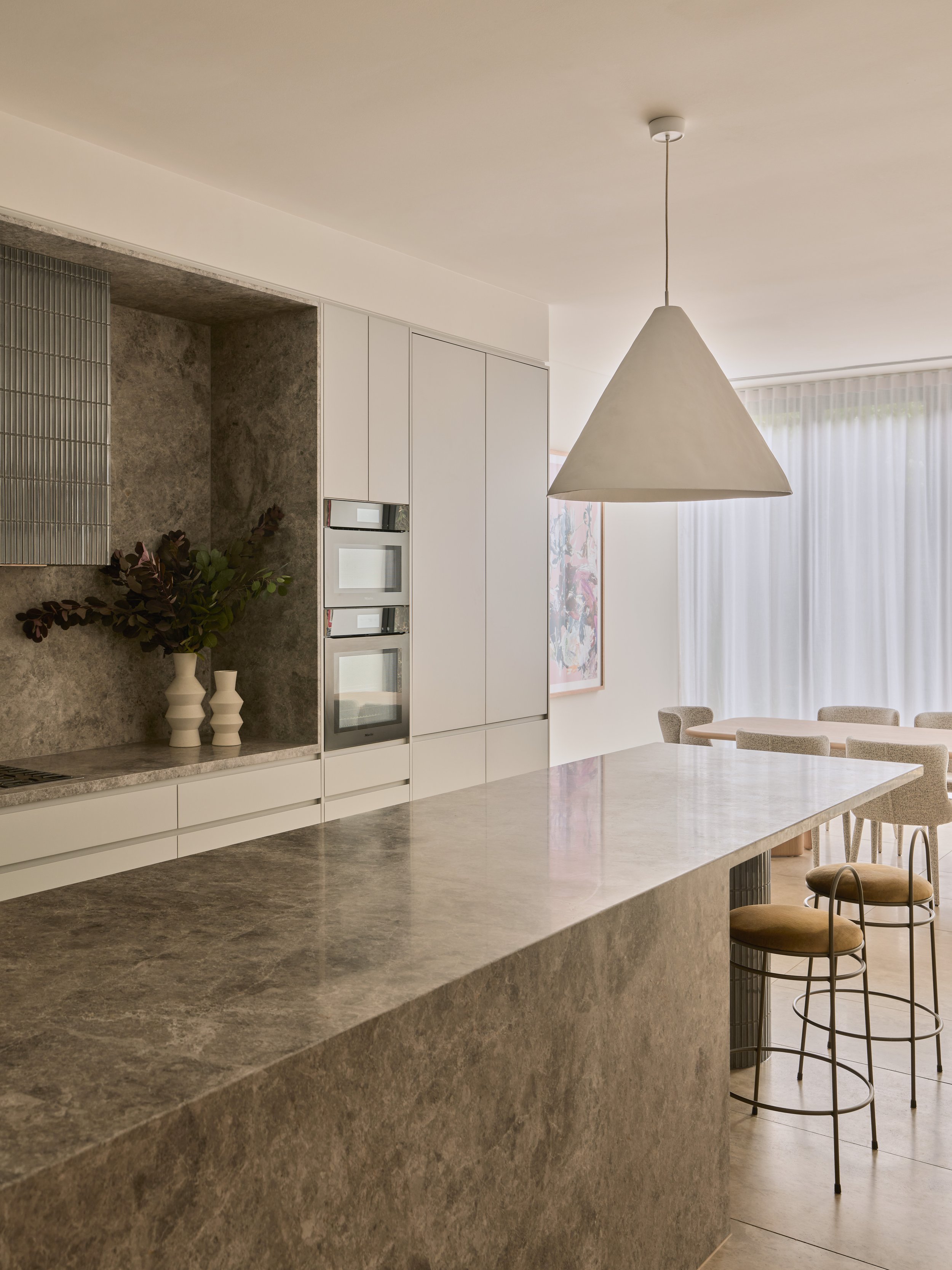 Modern kitchen with beige marble countertops, white cabinetry, and a white conical pendant light. There are three built-in ovens and decorative vases with greenery on the counter. In the background, there's a dining area with upholstered chairs and a