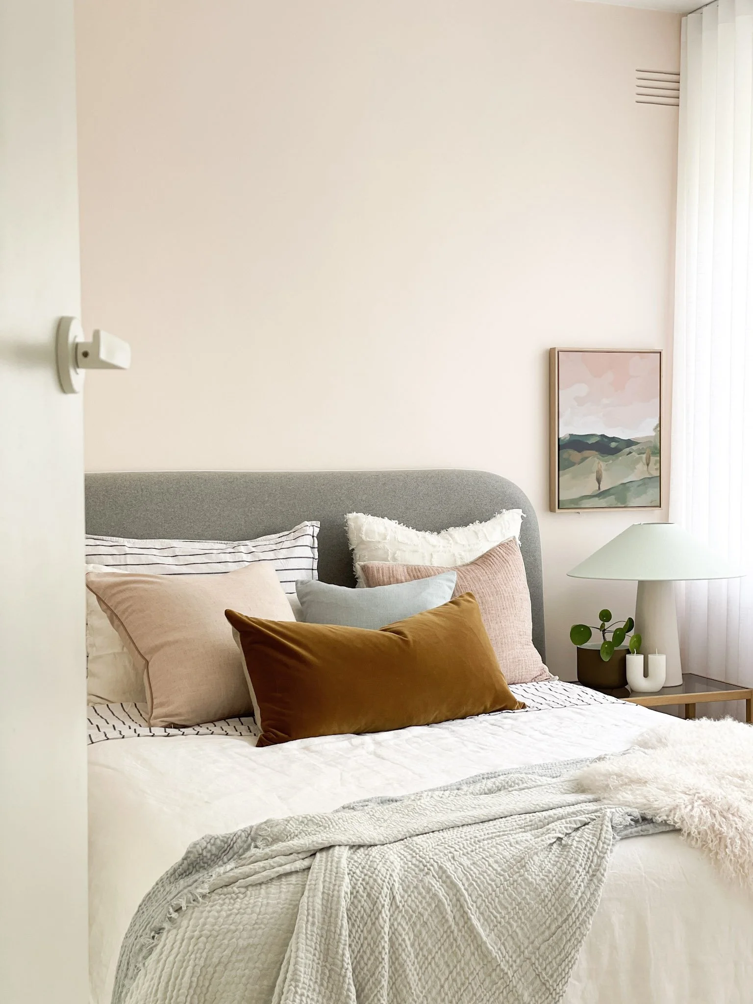 A neatly made bed with multiple pillows in soft colors, a cozy throw blanket, and a side table with a lamp and decorative plant, in a bright room with a pastel pink wall and a landscape painting.