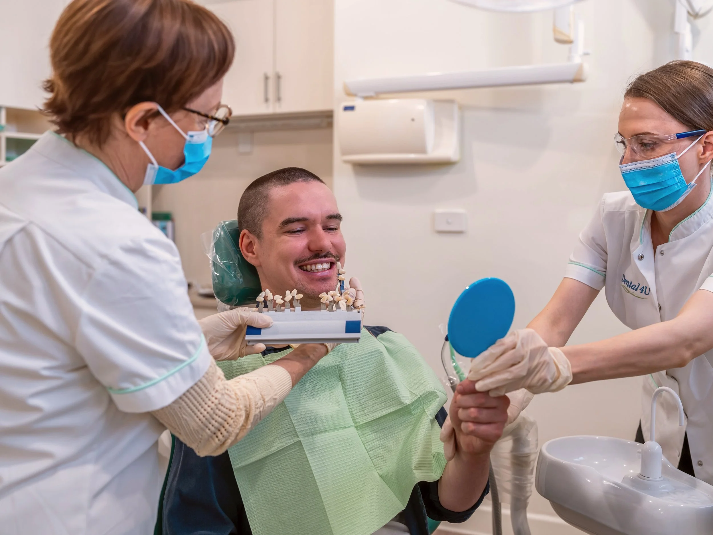 Male patient smiling at dental checkup while two female dental professionals, both wearing masks and gloves, show him a model of teeth and a mirror in a dental office.