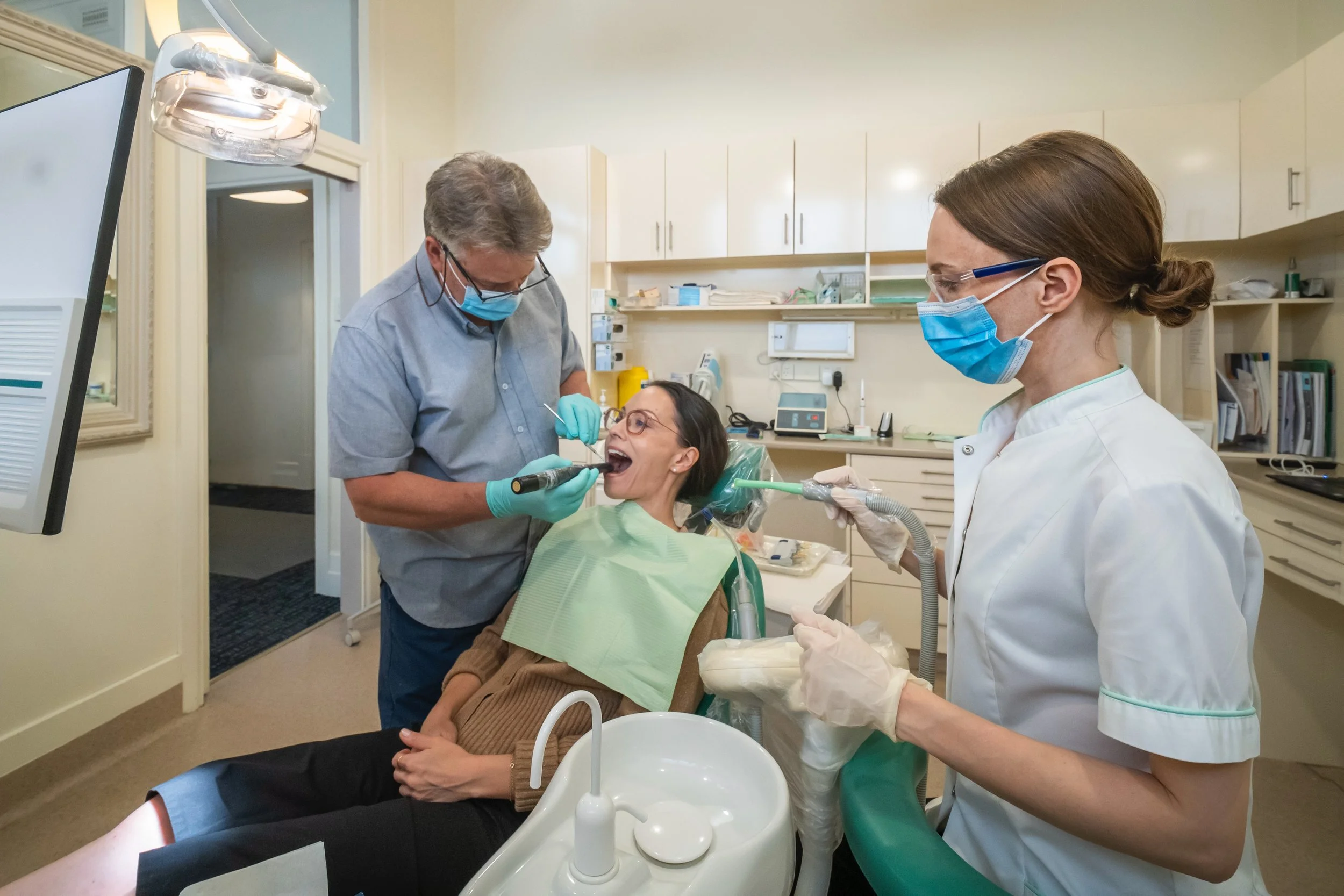 A woman at the dentist's office is receiving dental care from a male dentist and a female dental assistant, both wearing face masks, gloves, and dental scrubs, with the woman reclined in a dental chair.