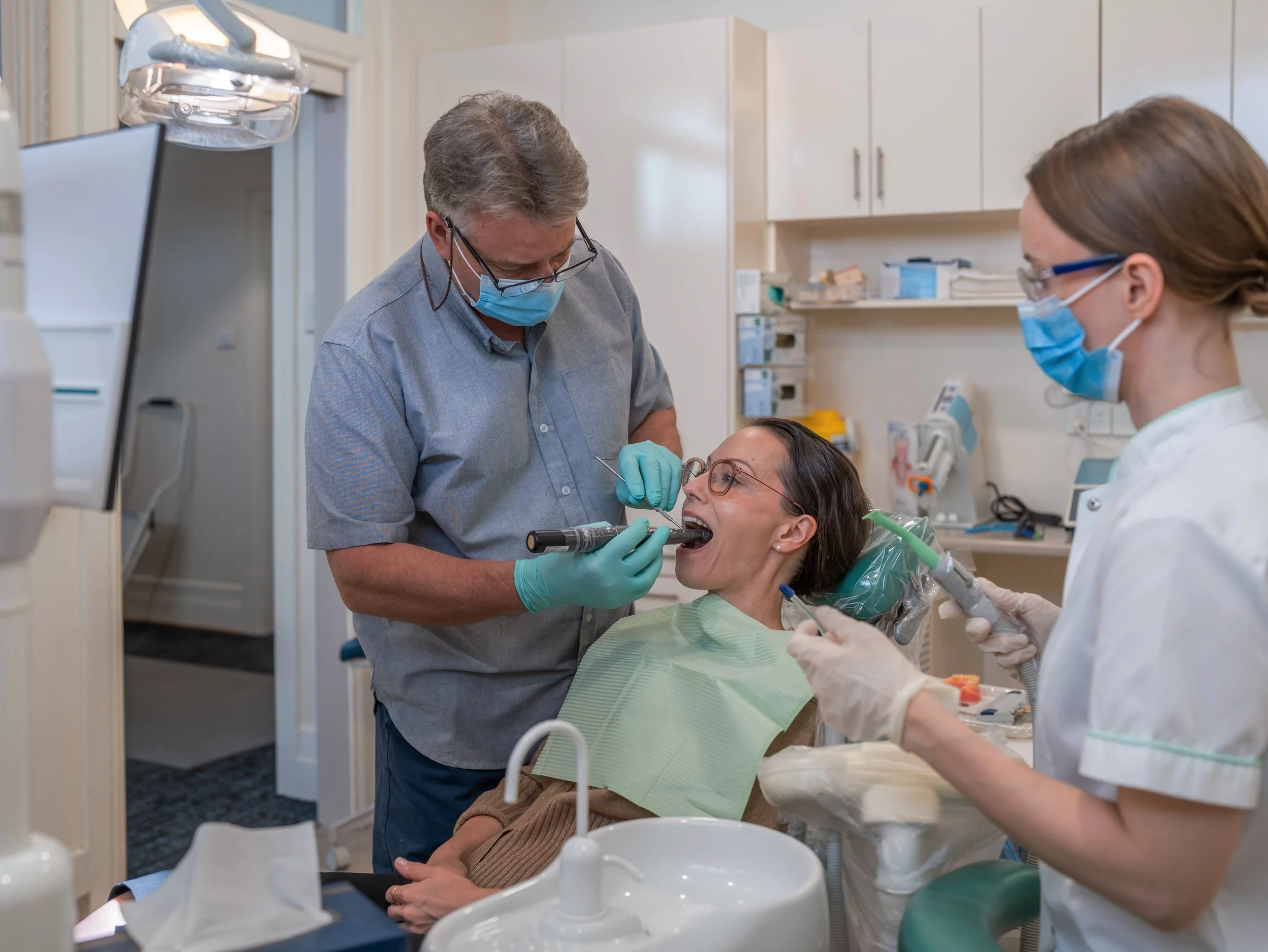 A dental professional  uses a dental scanner  to performs a dental examination or procedure on a female patient in a dental clinic, with another dental professional observing.