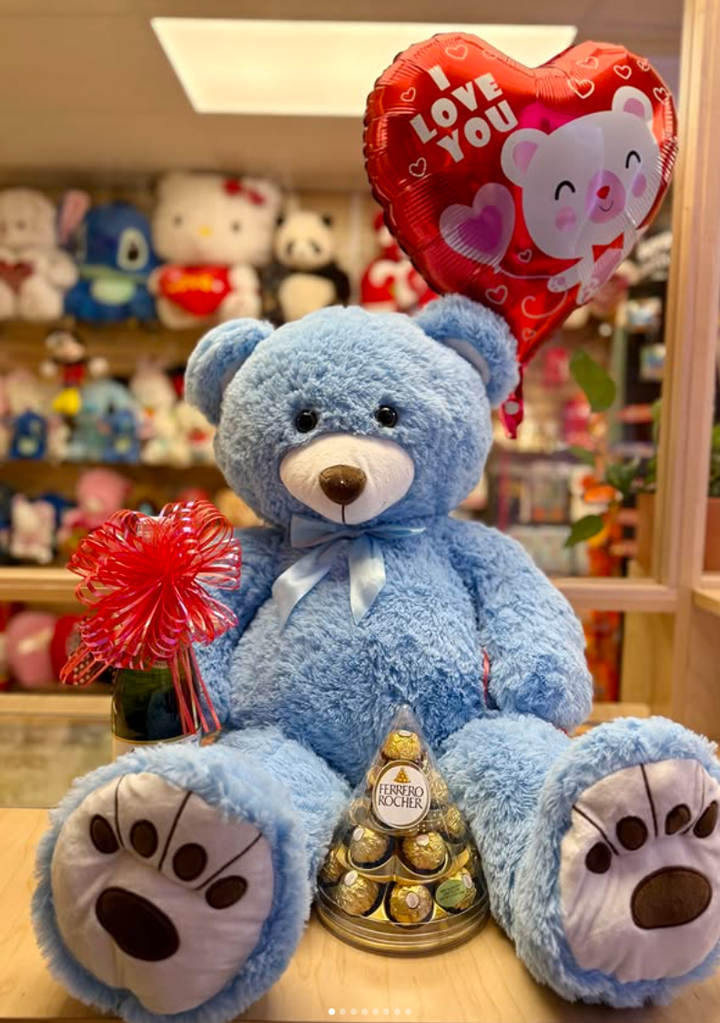 A blue teddy bear with a ribbon around its neck, holding a red gift item and a heart-shaped balloon that says "I love you" with a bear illustration, sitting in front of a display of stuffed animals, with Ferrero Rocher chocolates in a pyramid shape in front of it.