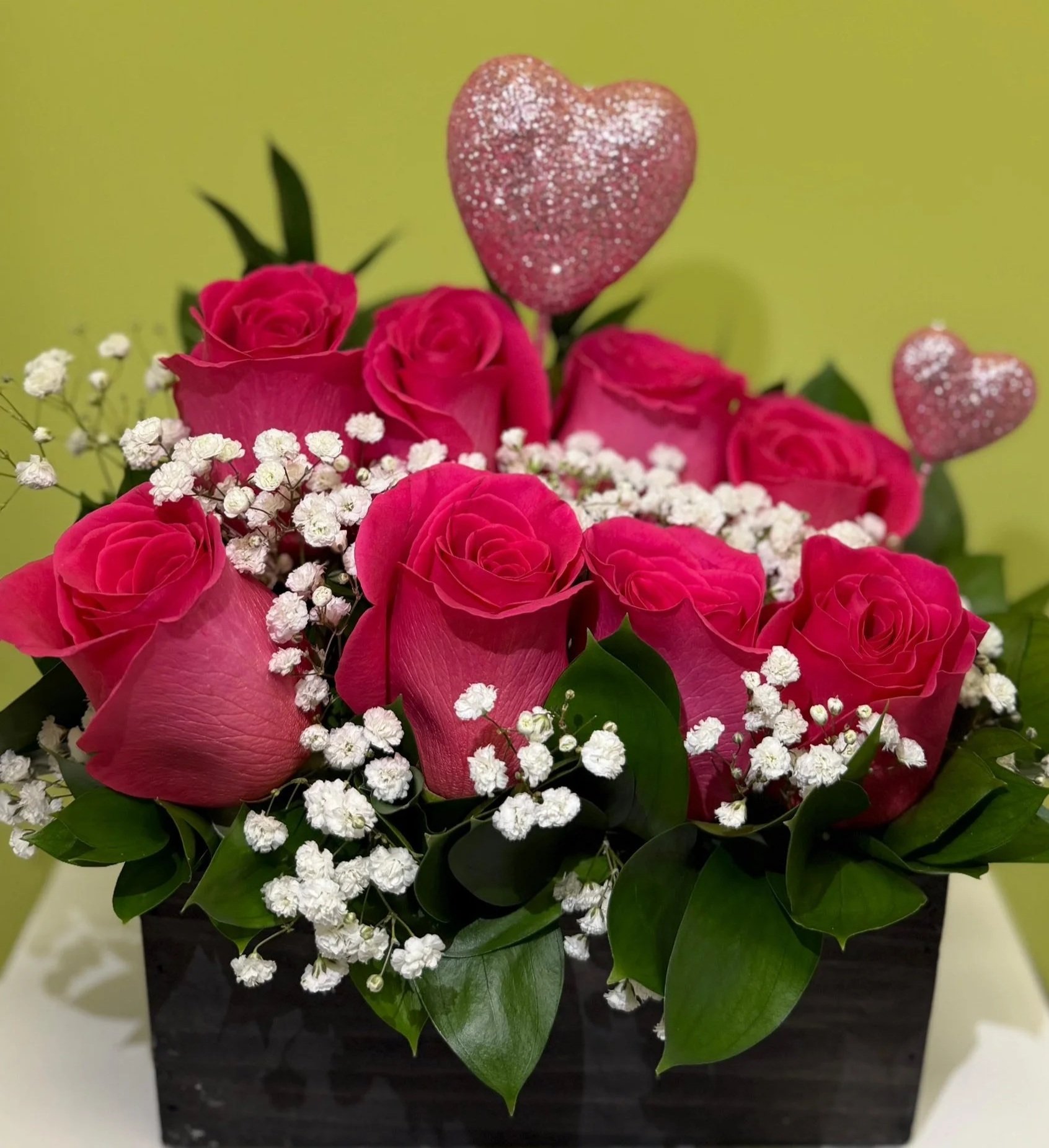 A flower arrangement of pink roses, baby's breath, and greenery in a black box, decorated with two glittery pink heart ornaments.