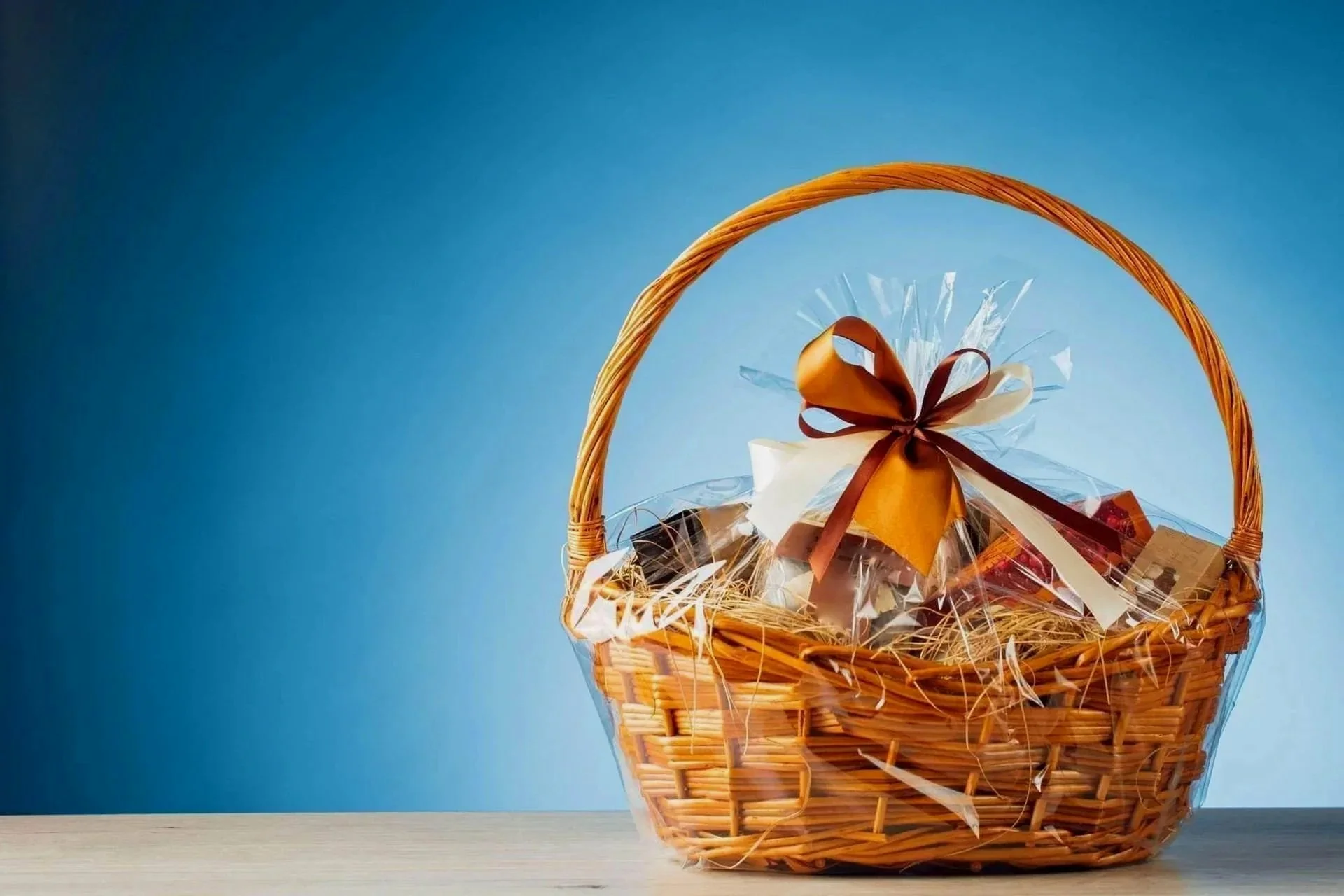 A wicker gift basket wrapped in clear cellophane with a large chocolate and cream-colored bow, sitting on a wooden surface against a blue background.