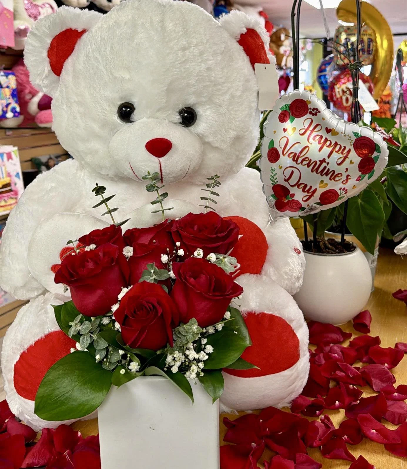 A stuffed white teddy bear with red patches on its ears and paws holding a bouquet of red roses, surrounded by rose petals on a table. There is a Valentine's Day balloon with red roses and gold hearts reading 'Happy Valentine’s Day' next to the bear, and a potted plant with green leaves in the background.