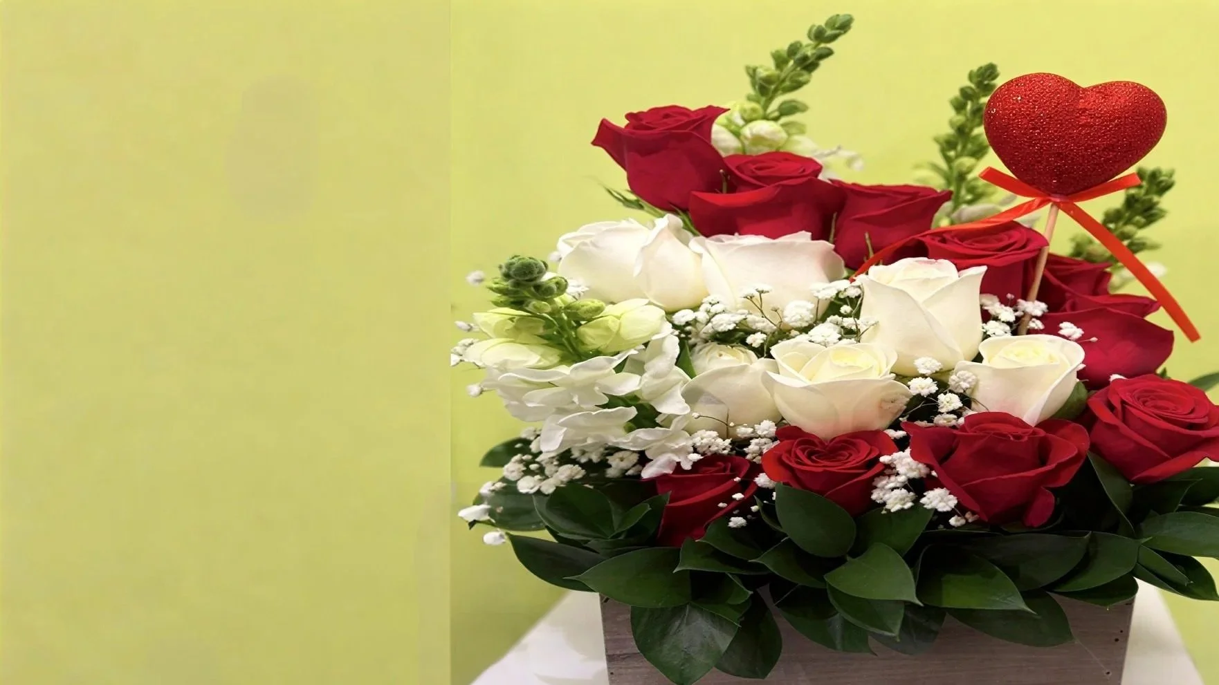 A floral arrangement with red and white roses, white baby's breath, and green foliage in a wooden box, with a red heart decoration on a stick.