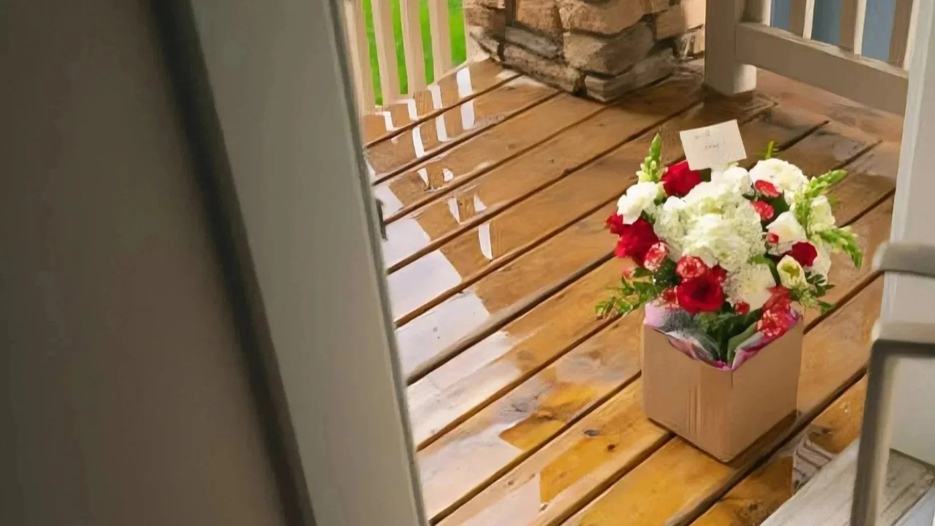 A bouquet of red and white flowers inside a beige paper wrap placed on a wooden deck outside a house.