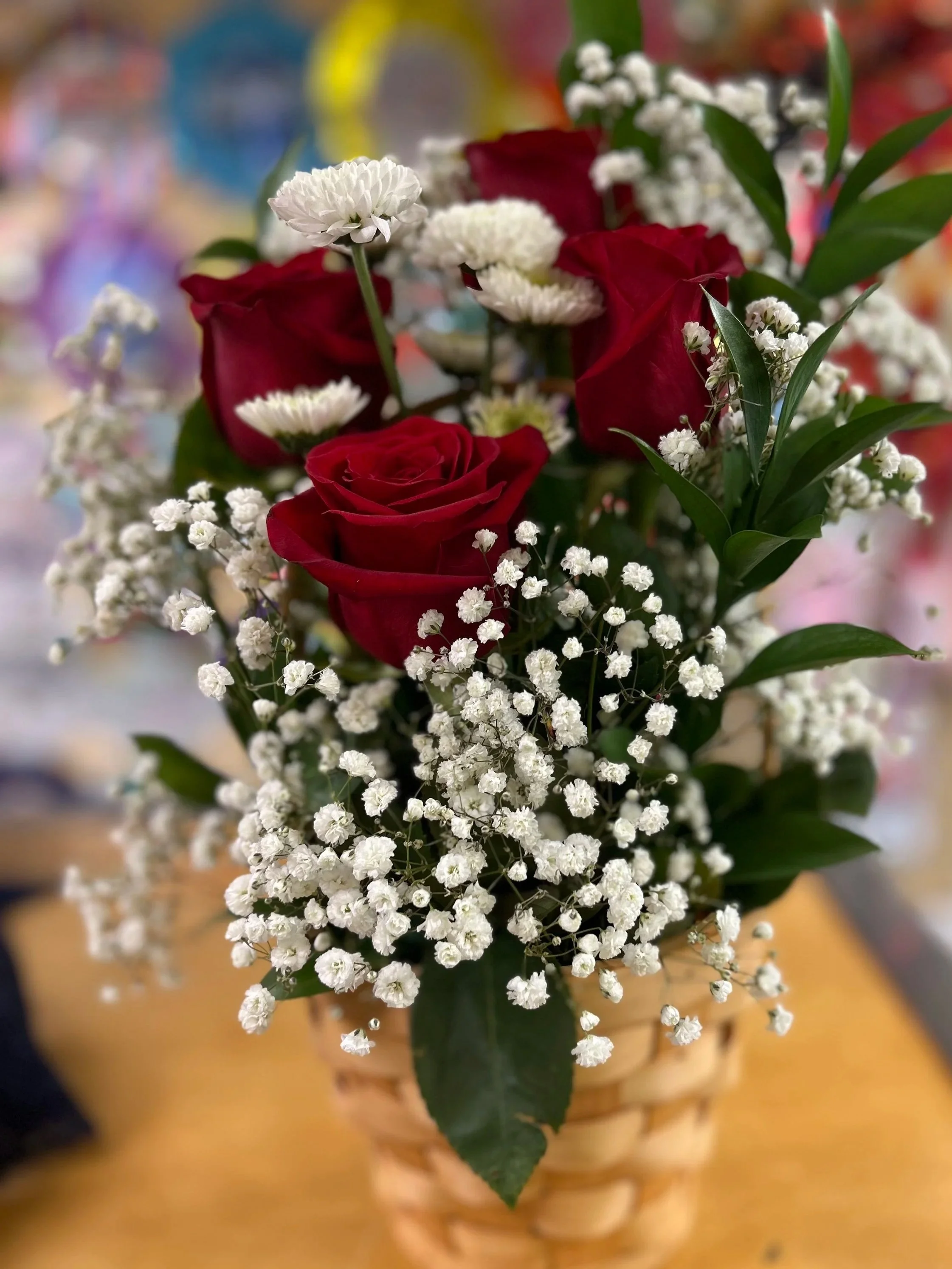 A bouquet of red roses, white baby's breath, and small white and purple flowers in a woven basket.