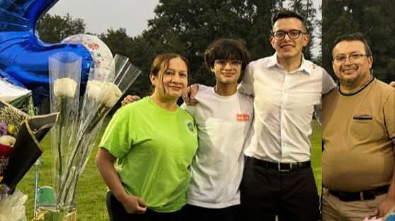 Four people standing outdoors in a park, smiling, with a backdrop of trees, and balloons nearby.