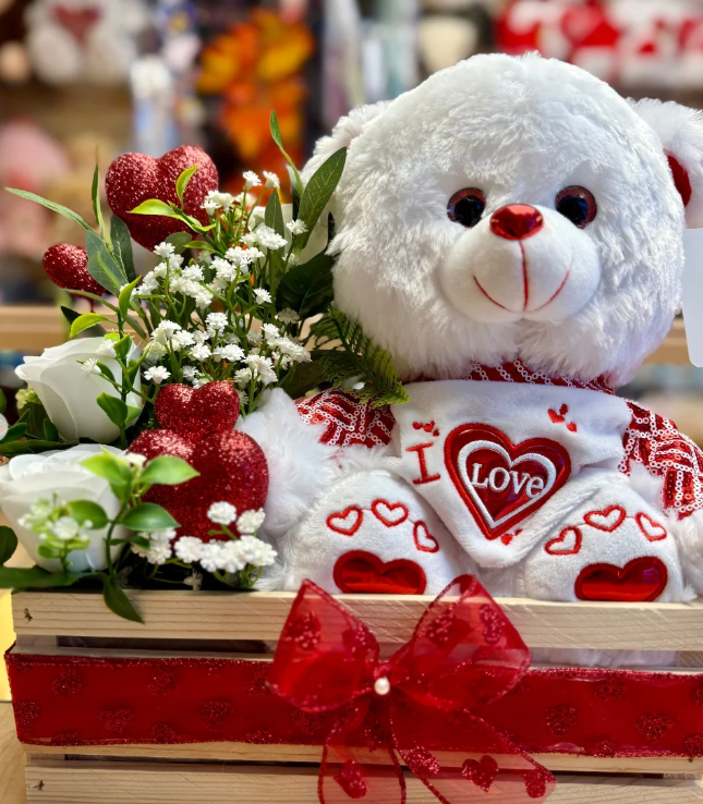 A white teddy bear with red accents, holding a heart-shaped sign that says "LOVE," surrounded by red and white Valentine's Day decorations, artificial red glitter hearts, white roses, small white flowers, and green leaves in a wooden basket decorated with a red ribbon.