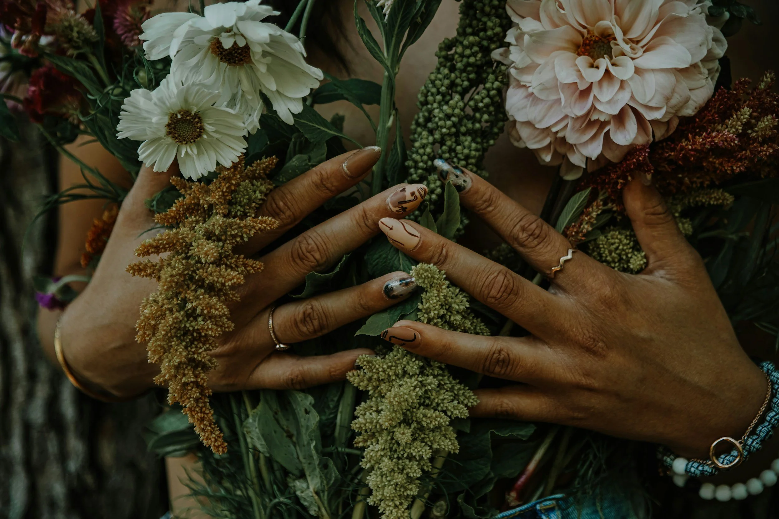 Two hands with manicured nails and jewelry holding a bouquet of mixed flowers, including white daisies, pale pink dahlias, and green foliage.